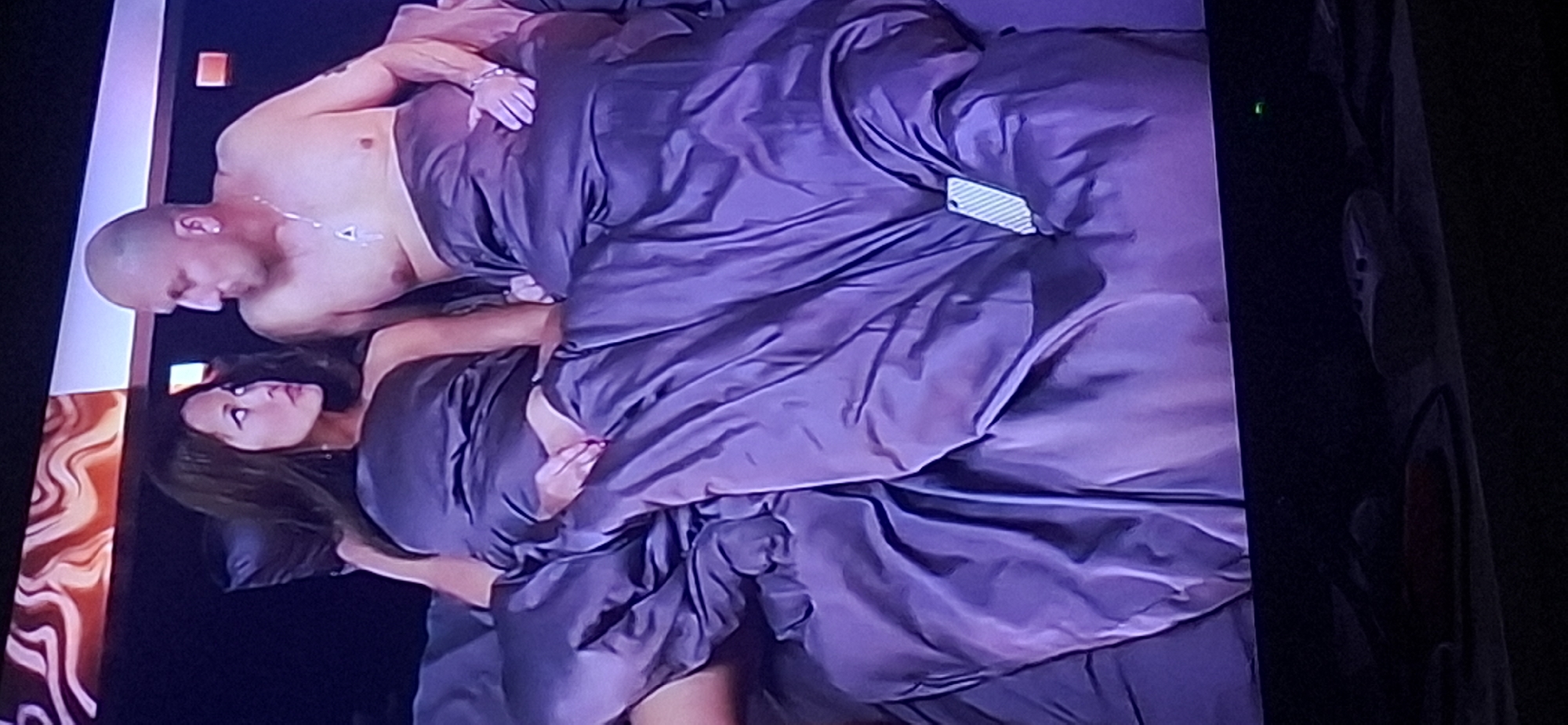 The image depicts a man and a woman lying in bed, partially covered by a dark purple duvet. The woman, with dark hair, is looking to her left, while the bald man is looking towards her. Both are wearing metallic-looking bottoms. The man has a necklace with a pendant and is wearing earrings. The background shows a patterned headboard and a dark wall. The image appears to be a screenshot from a television screen, as evidenced by the screen's edge and a small green light in the lower right corner, possibly an indicator light. There are also objects on a shelf below the screen, including what appears to be a remote control. The scene is not identifiable as being in Yerocham, Israel.