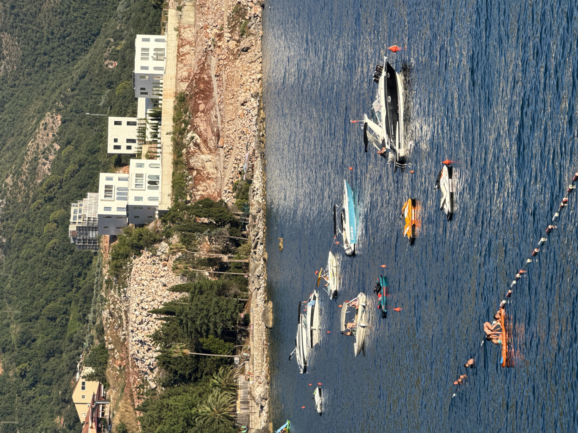 This is a high-angle, medium-shot photograph of the coastline in Kamenovo, Montenegro. The scene is dominated by the deep blue water of the Adriatic Sea, with several boats of various sizes scattered across it. In the foreground, two people are positioned on an orange inflatable raft, with one person appearing to be sitting while the other is leaning over them. A series of buoys and a rope mark a boundary in the water between the inflatable raft and the shore.
Further out on the water, there are several other vessels, including speedboats, a jet ski, and a smaller boat with a canopy. Some of these boats appear to be occupied by individuals.
The background of the image features a rocky, red-hued cliff face that rises steeply from the coastline. Atop this cliff, a cluster of modern, white buildings with geometric designs are visible, contrasting with the natural landscape. Lush green trees and vegetation are also present on the slopes of the hill behind the buildings. On the lower left side of the frame, a section of the shore is visible, with a beach area, umbrellas, and more people enjoying the seaside. The sky is clear, indicating a sunny day. The overall impression is one of a popular tourist destination with recreational activities taking place on the water.