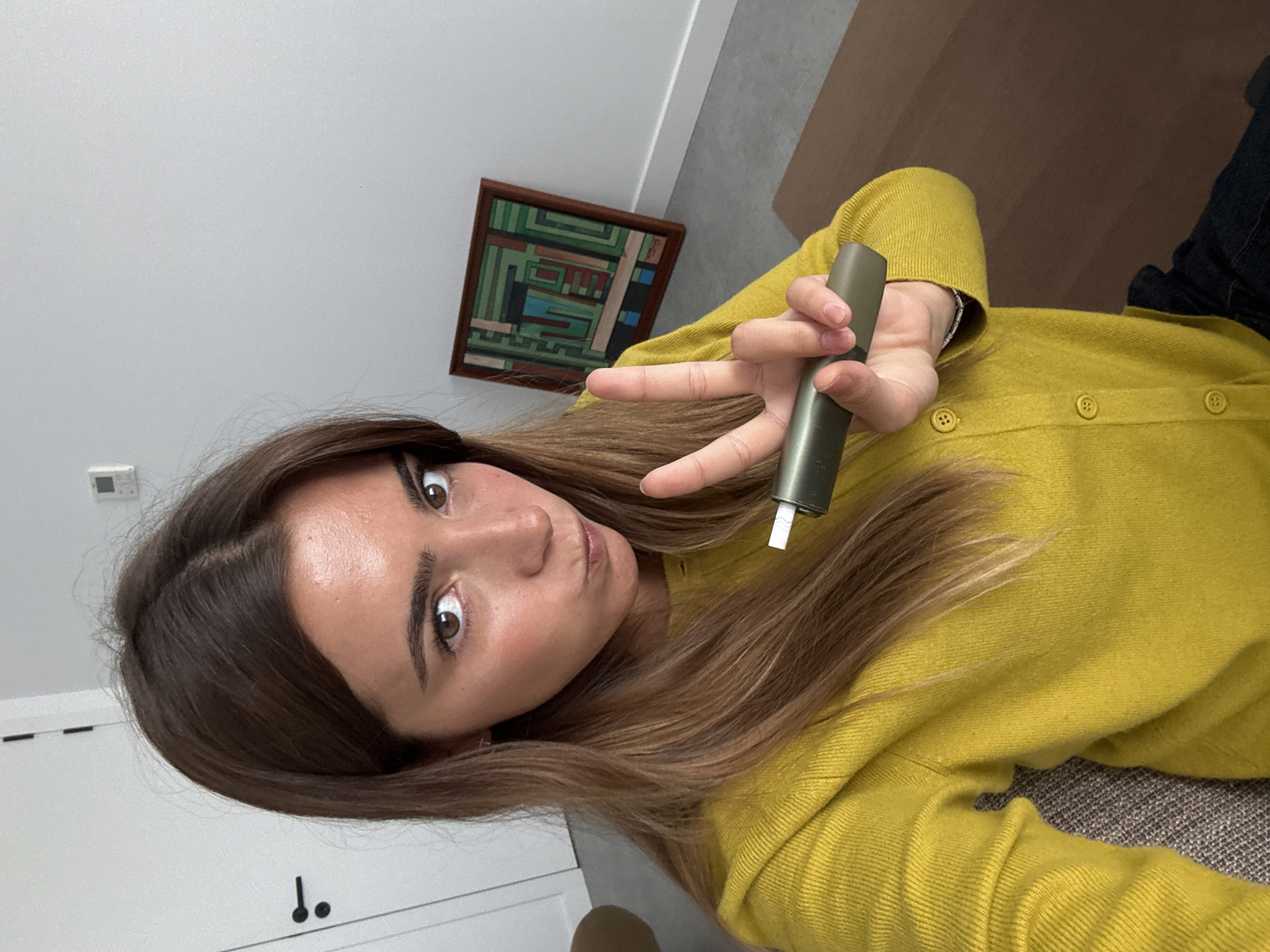 A young woman poses for a selfie indoors. She is fair-skinned with brown eyes and shoulder-length brown hair with blonde highlights. She wears a yellow cardigan with buttons. She holds a gray electronic device in her right hand and makes a peace sign. The setting appears to be a modern home. A framed piece of abstract art hangs on the white wall behind her. A white door with a black handle is visible in the lower left. There is a gray floor and a brown surface in the upper right. The location is not apparent from this image.