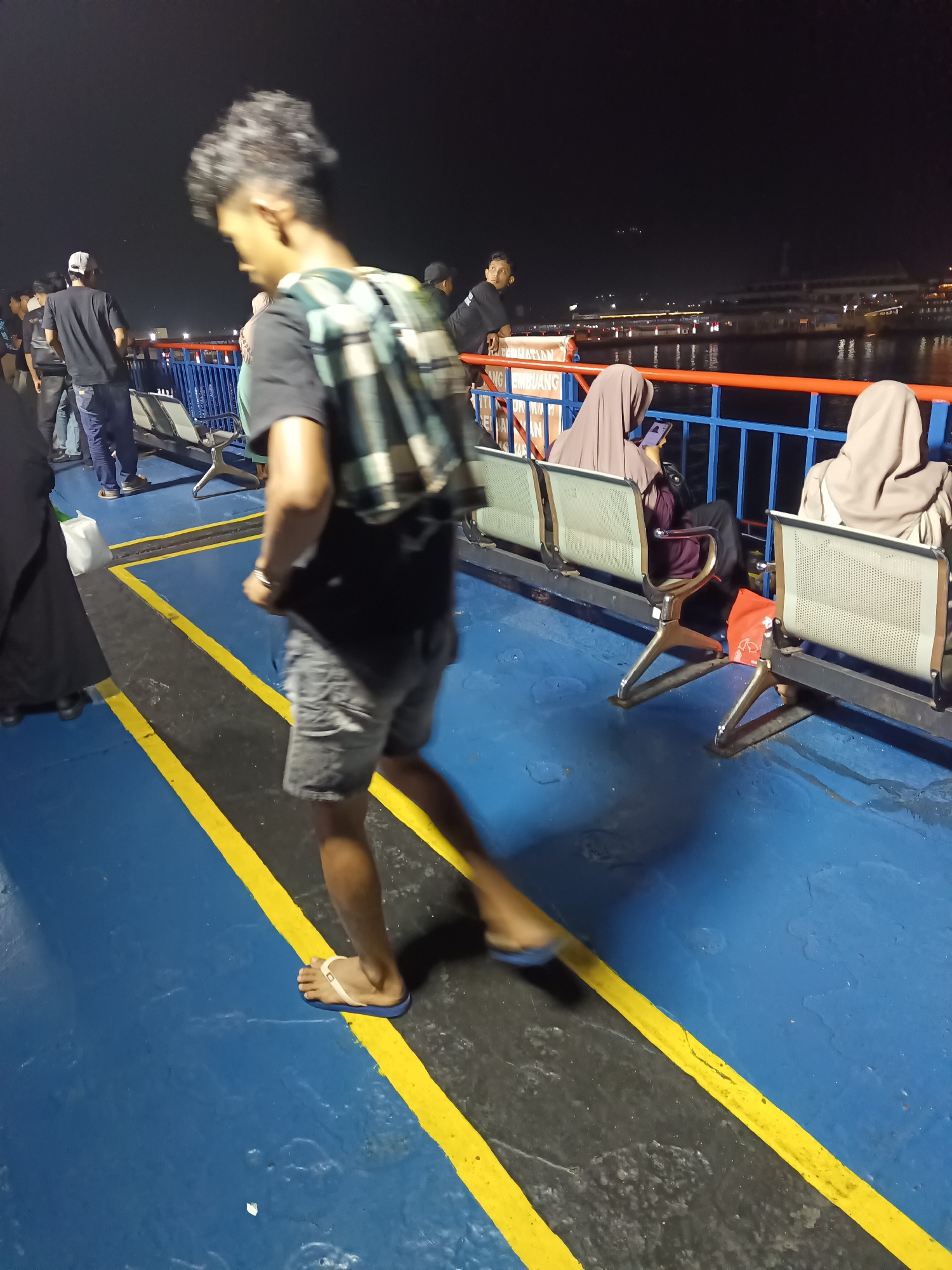 This image depicts a nighttime scene on what appears to be a ferry in Cilegon, Indonesia, likely near the port. In the foreground, a young person with curly hair is walking away from the camera, wearing a black t-shirt, grey shorts, and flip-flops. They are carrying a checkered backpack. The deck of the ferry is painted blue with yellow and black striped lines marking a walkway.
Further back, several other individuals are visible. To the left, a group of people are standing, including one person wearing a black t-shirt and a white cap, and another in a black jacket. On the right side, three people are seated on a row of blue and grey chairs. Two of these individuals are wearing hijabs; one in a purple hijab is looking at their phone, and the other in a light brown hijab is also seated. A bright orange bag is visible on the deck near the seated individuals.
In the background, a body of water is visible, with lights from a distant city or port illuminating the night sky. A large building or structure is dimly lit across the water. The overall environment suggests a functional, open-air area on a mode of transport, likely a ferry, at night.