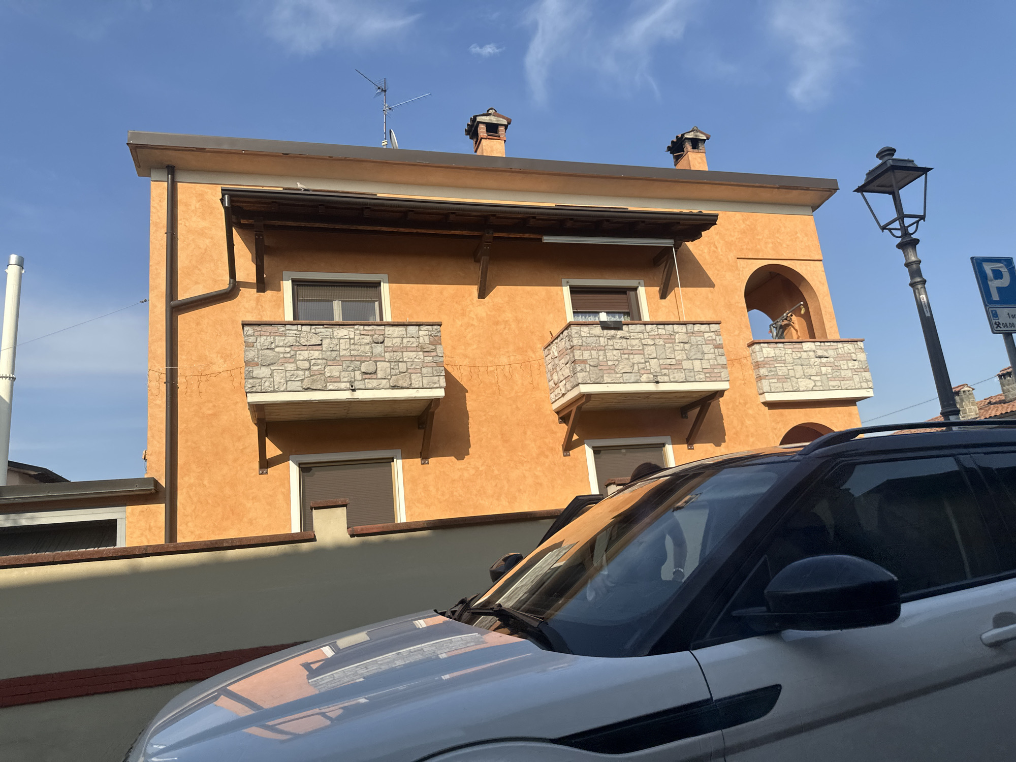 In Rudiano, Italy, a silver SUV is parked on a street in front of an orange building with stone accents. The building has two balconies with stone railings and windows with brown roller shutters. The roof is visible with two chimneys and an antenna. A black lamppost stands to the right of the building, and a blue parking sign is mounted on it. The sky is clear blue with a few wispy clouds. The image captures a sunny day in the town.