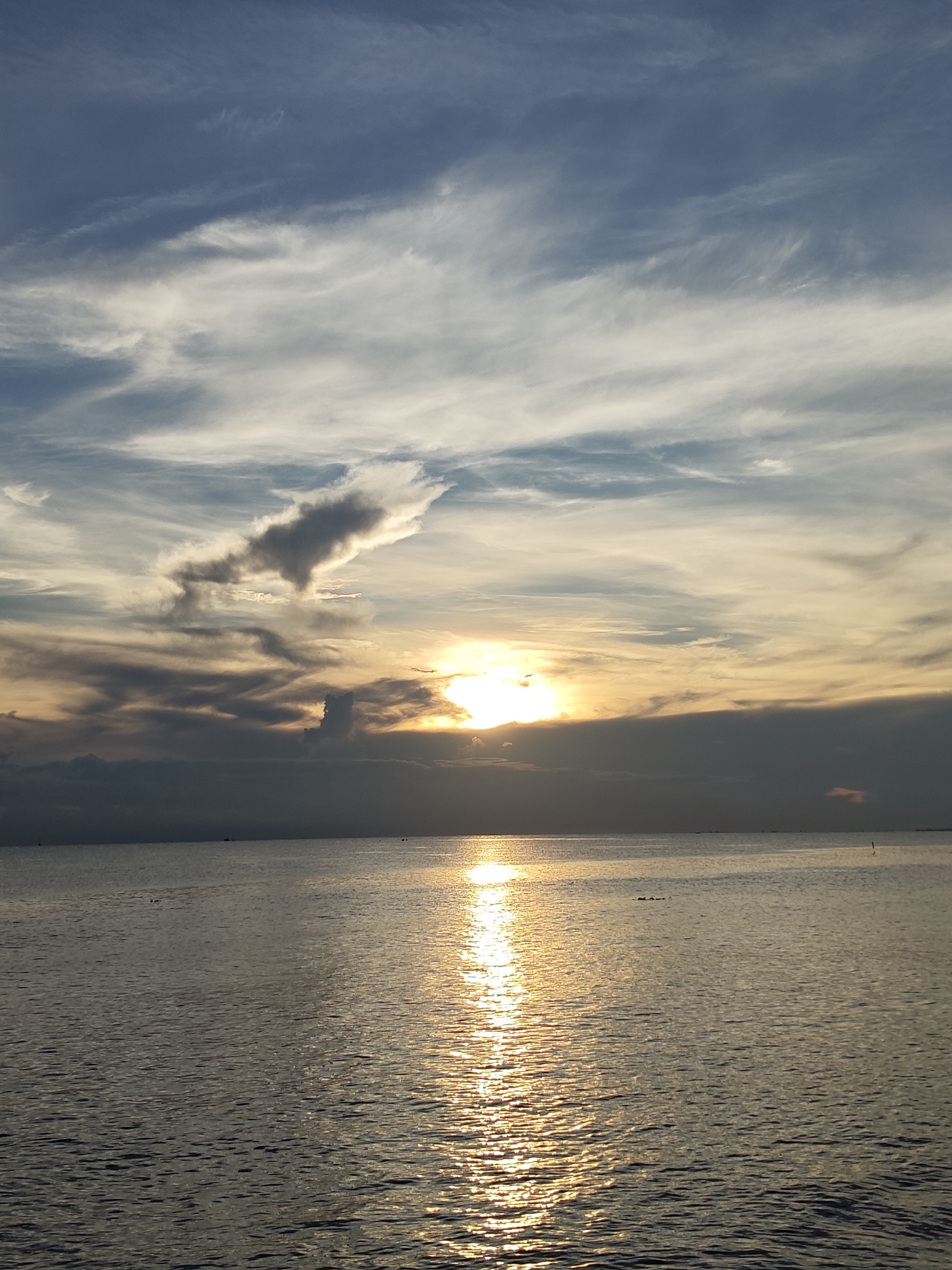 The image shows a sunset over the sea, possibly taken in Lhokseumawe, Indonesia. The sun is partially obscured by clouds, casting a bright reflection on the water. The sky is a mix of blue, white, and golden hues, with various cloud formations. The sea is relatively calm, with a few ripples reflecting the sunlight. There are no visible people or objects on the shore, and the scene appears peaceful and serene.