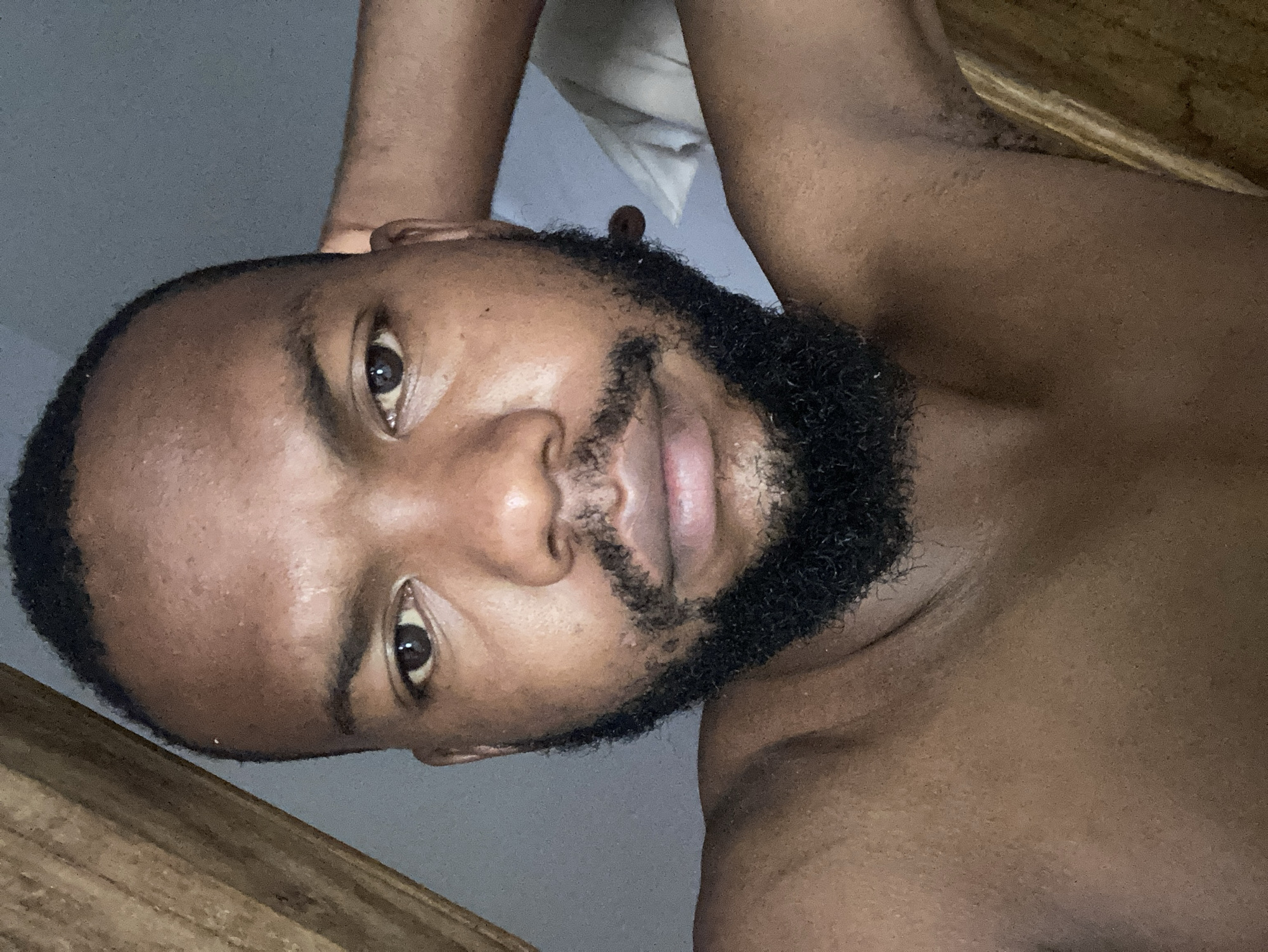 A close-up, rotated portrait shows a Black man with a dark, tightly curled beard and short hair. His skin tone is brown. He is topless. Part of a wooden surface appears in the lower left and upper right corners. The background is indistinct, appearing to be a light gray wall. The photograph's location is not provided.