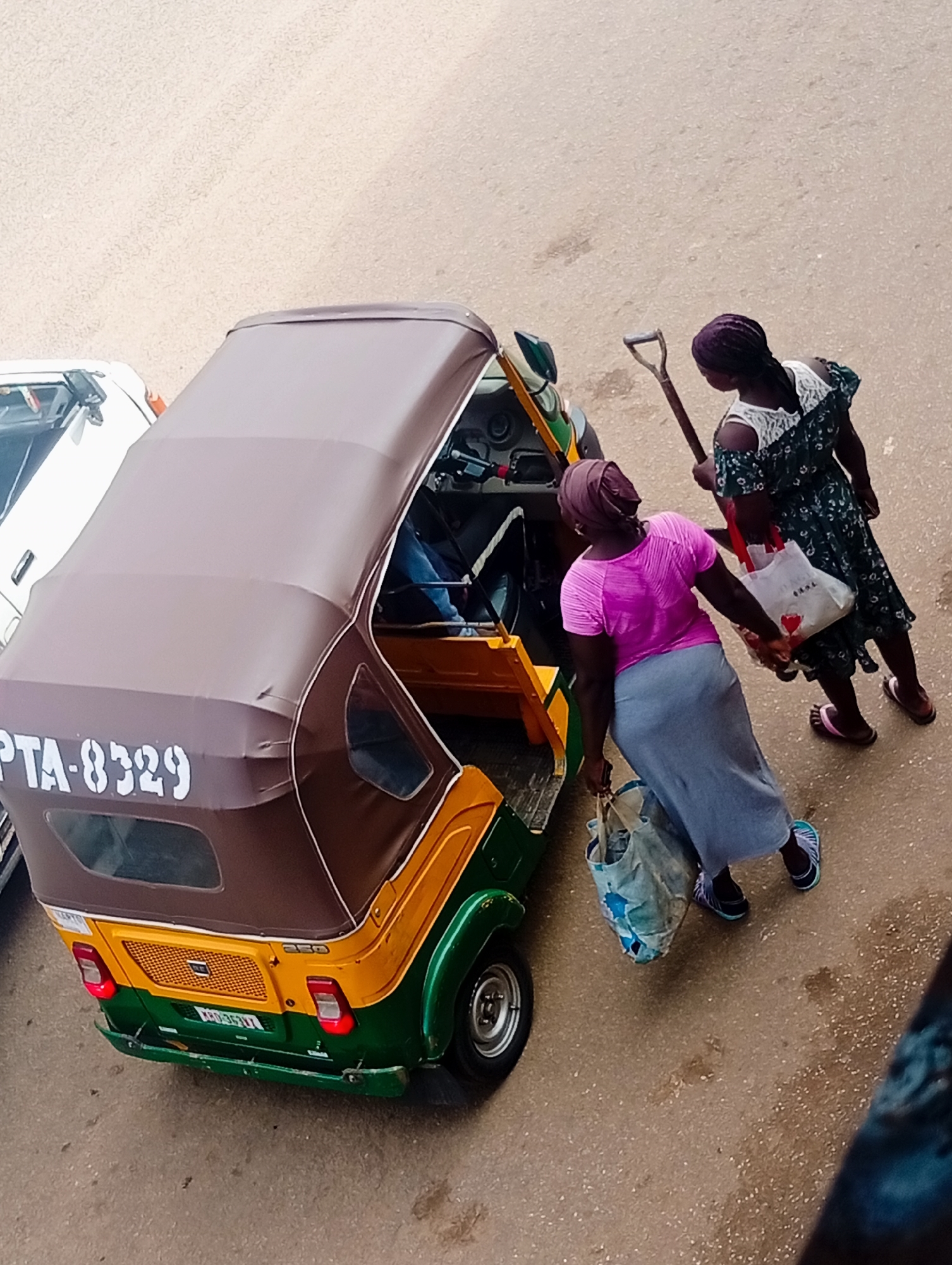 An overhead view captures two women and a three-wheeled auto-rickshaw on a dusty, light brown road in Bukuru, Nigeria. The auto-rickshaw is positioned in the foreground, facing right. It has a brown canopy, a yellow front, and green sides. The license plate reads "PTA-8329".
One woman, on the left, wears a pink t-shirt and a long grey skirt. She is carrying a light blue and white patterned bag and appears to be walking away from the auto-rickshaw.
The second woman, on the right, wears a dark green floral print dress with white lace trim on the shoulders. She holds a long-handled tool, possibly a shovel, with a D-shaped grip, in her right hand. In her left hand, she carries a white shopping bag with a red heart design. She is also holding a light colored shopping bag by its handles. This woman is positioned behind and slightly to the right of the first woman, and they appear to be moving together. Her feet are visible, wearing brown sandals. The first woman is wearing blue and white striped sandals.
A white vehicle is partially visible on the left side of the frame, to the left of the auto-rickshaw. The road surface shows some dark wet patches, suggesting recent moisture. The background consists of more of the dusty road.