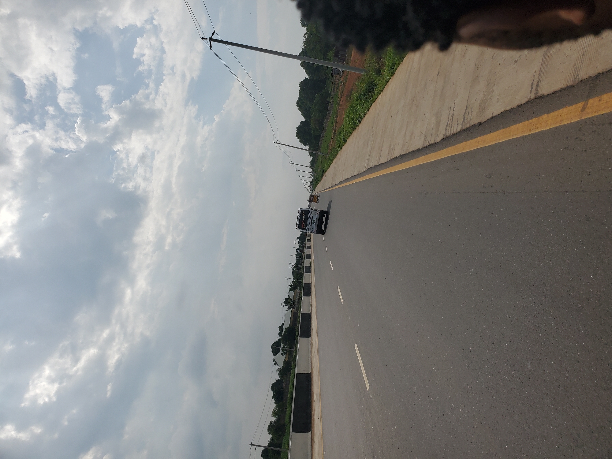 The image shows a road with a clear sky overhead. The road has white dashed lines marking the lanes. There is a concrete sidewalk on the right side of the road with a yellow line painted along its edge. A few vehicles are visible on the road, including a white van and a yellow vehicle. Trees and buildings can be seen in the distance. Power lines stretch across the scene, supported by utility poles. The image appears to be taken from a moving vehicle, as indicated by the slight blur and the perspective. The location is not the city of Winters, United States of America.