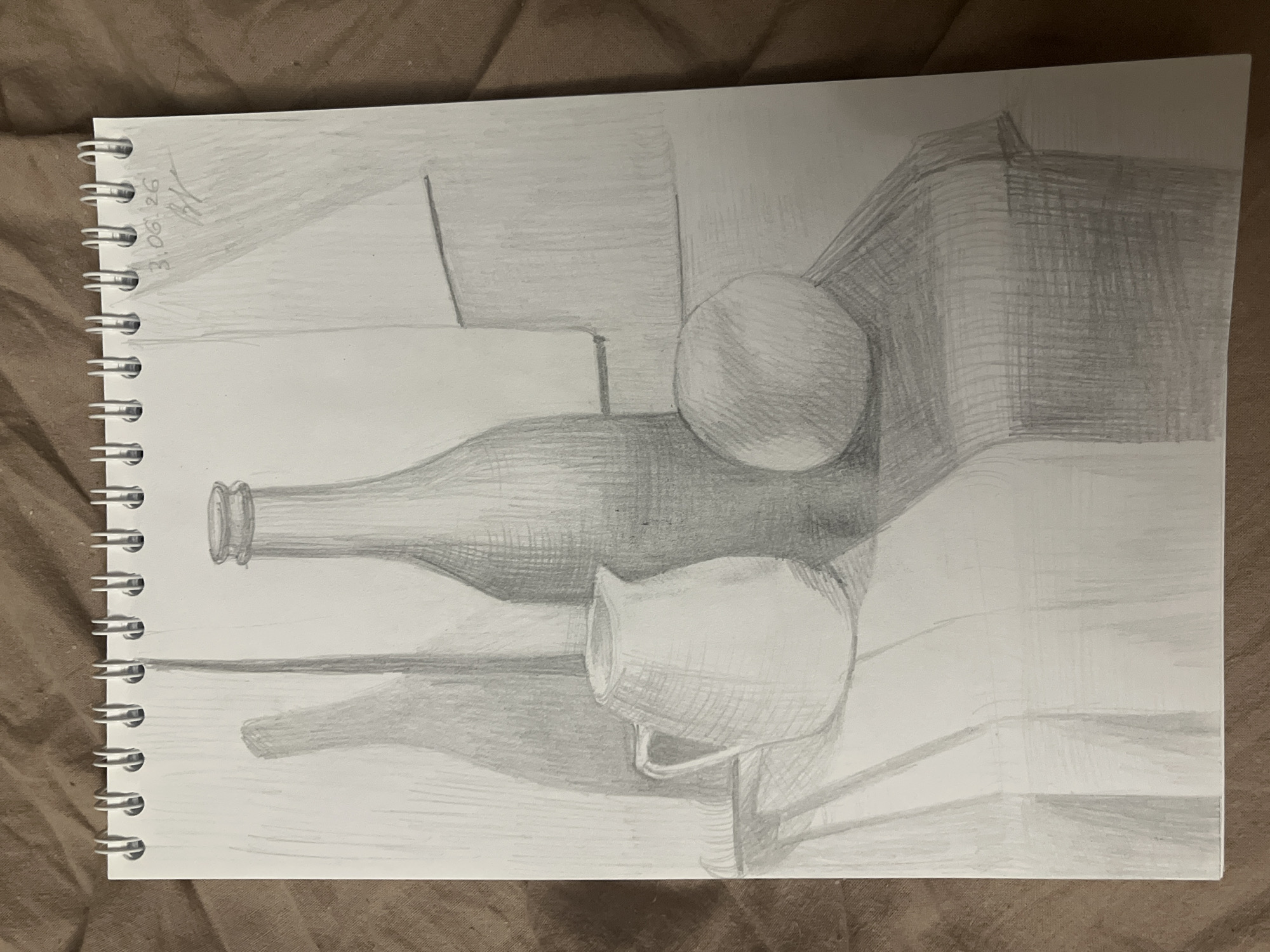 A pencil drawing depicts a still life composition rendered in grayscale tones. The primary subjects include a bottle lying horizontally in the mid-ground, a spherical object (resembling an orange or apple) positioned above and slightly behind the bottle, and a small jug or pitcher with a handle located in front of and to the right of the bottle.
The objects are arranged on a flat surface, with a distinct background plane behind them, suggesting a table against a wall. The drawing demonstrates an academic study of form, light, and shadow. Shading is achieved through hatching and cross-hatching techniques, giving volume to the objects and defining the planes of the background and foreground. Shadows are cast to the right of the objects, indicating a light source originating from the upper left.
Notable details include the texture created by the pencil strokes, particularly on the spherical object, the jug, and the background wall. The drawing is contained within a spiral-bound sketchpad. Faint pencil markings in the top-left corner read "3.08.26," likely indicating a date, and possibly artist's initials such as "HK" or similar. No activity is taking place within the depicted scene; it is a static arrangement of inanimate objects.