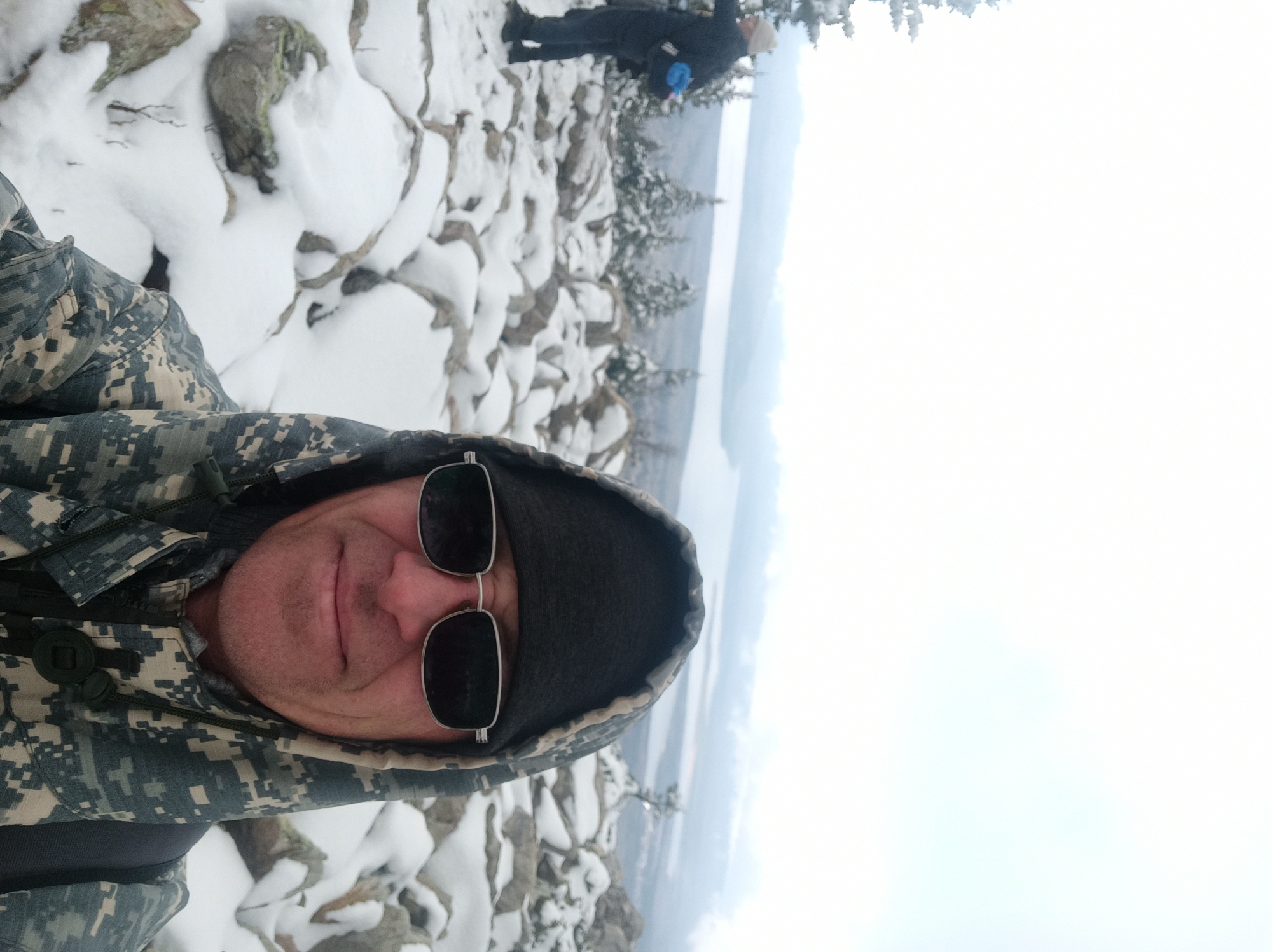 A person wearing sunglasses and a camouflage hooded jacket is taking a selfie. The background shows a snowy, rocky terrain with some trees and a body of water. Another person is visible further back in the scene. The location appears to be a mountainous area, likely in winter, based on the snow cover. Given the prompt's reference to Ziuratkul', Russia, this is likely the location, though further details are needed to pinpoint the exact spot within the city.