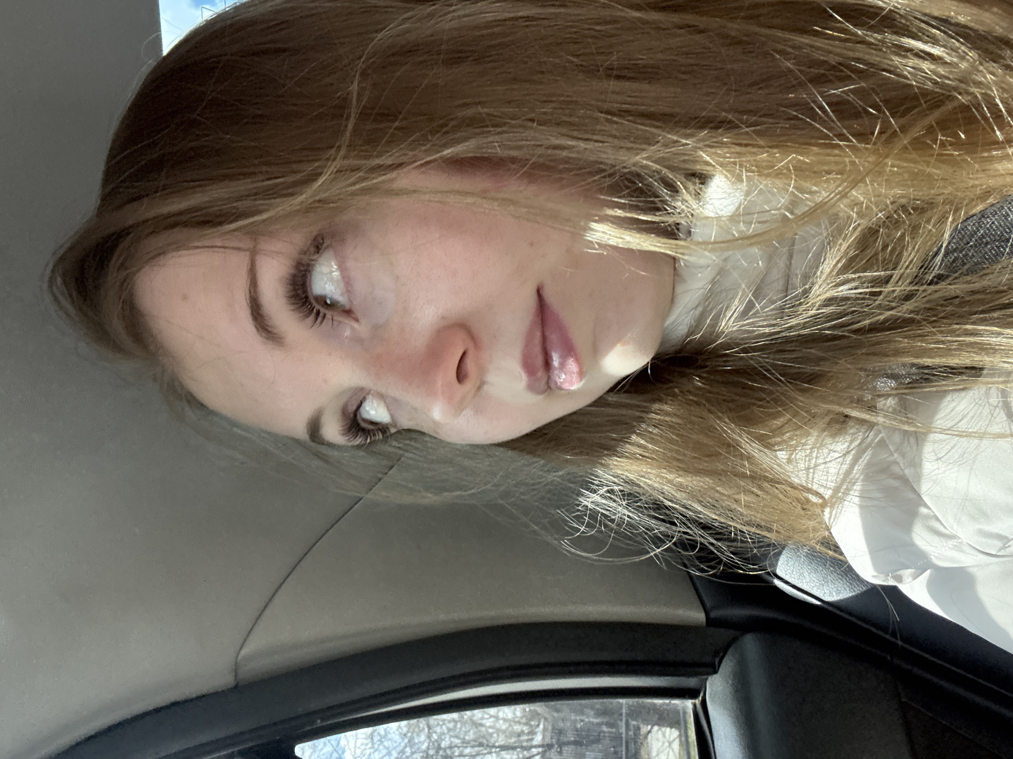 A close-up portrait shows a young person, likely female, inside what appears to be a car. The image is oriented with the person's head significantly tilted, almost sideways, suggesting she is looking upwards or to the side, and the photo itself is rotated 90 degrees counter-clockwise.
The subject has long, flowing light brown hair with golden highlights, some strands falling across her face. Her left eye is visible, accentuated by long, dark eyelashes and a streak of white eyeliner on the lower lash line. Her lips are slightly parted and appear glossy. She is wearing a white top, possibly a turtleneck or a collared shirt, and a dark strap, resembling a seatbelt or bag strap, is visible near her shoulder.
The background consists of the car's light gray headliner and a dark window frame. Through the window, a blurred outdoor scene with bare tree branches against a bright sky is visible, indicating daytime. Bright sunlight illuminates the person's hair and face. The overall expression is relaxed and somewhat contemplative. No visible text is present in the image.