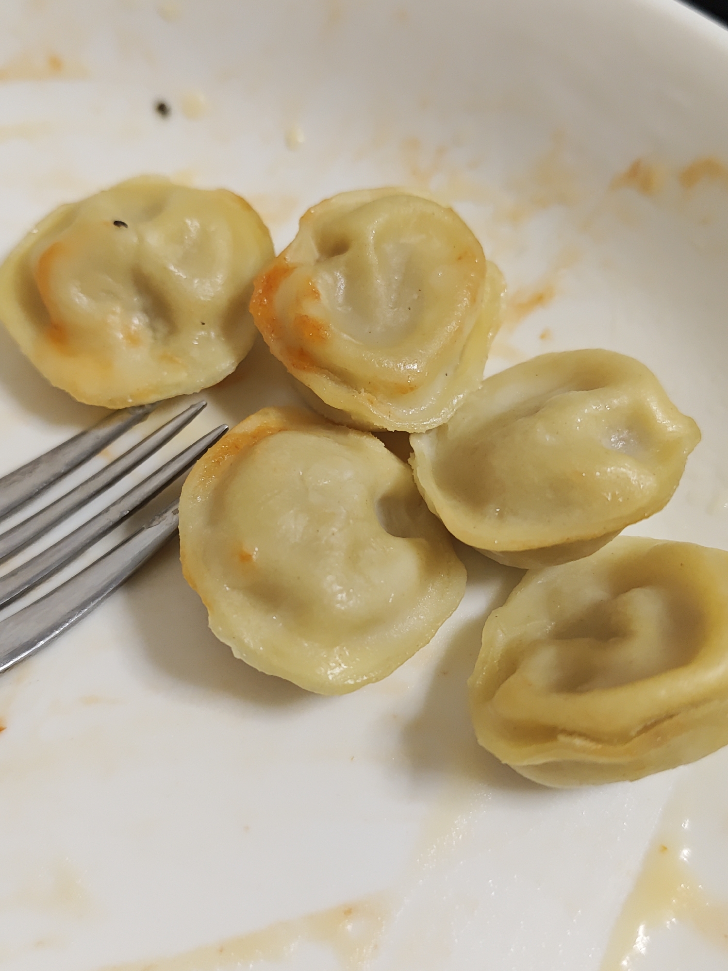 The image shows a close-up of five cooked dumplings on a white plate. A fork is visible to the left of the dumplings. The dumplings appear to have a light orange hue, possibly from a sauce or seasoning. There are some small dark specks on the dumplings and the plate, some of which appear to be insects, specifically fruit flies. The plate has remnants of what looks like a reddish-orange sauce. The lighting suggests an indoor setting. There is no indication of the city of Saint Petersburg, Russia.