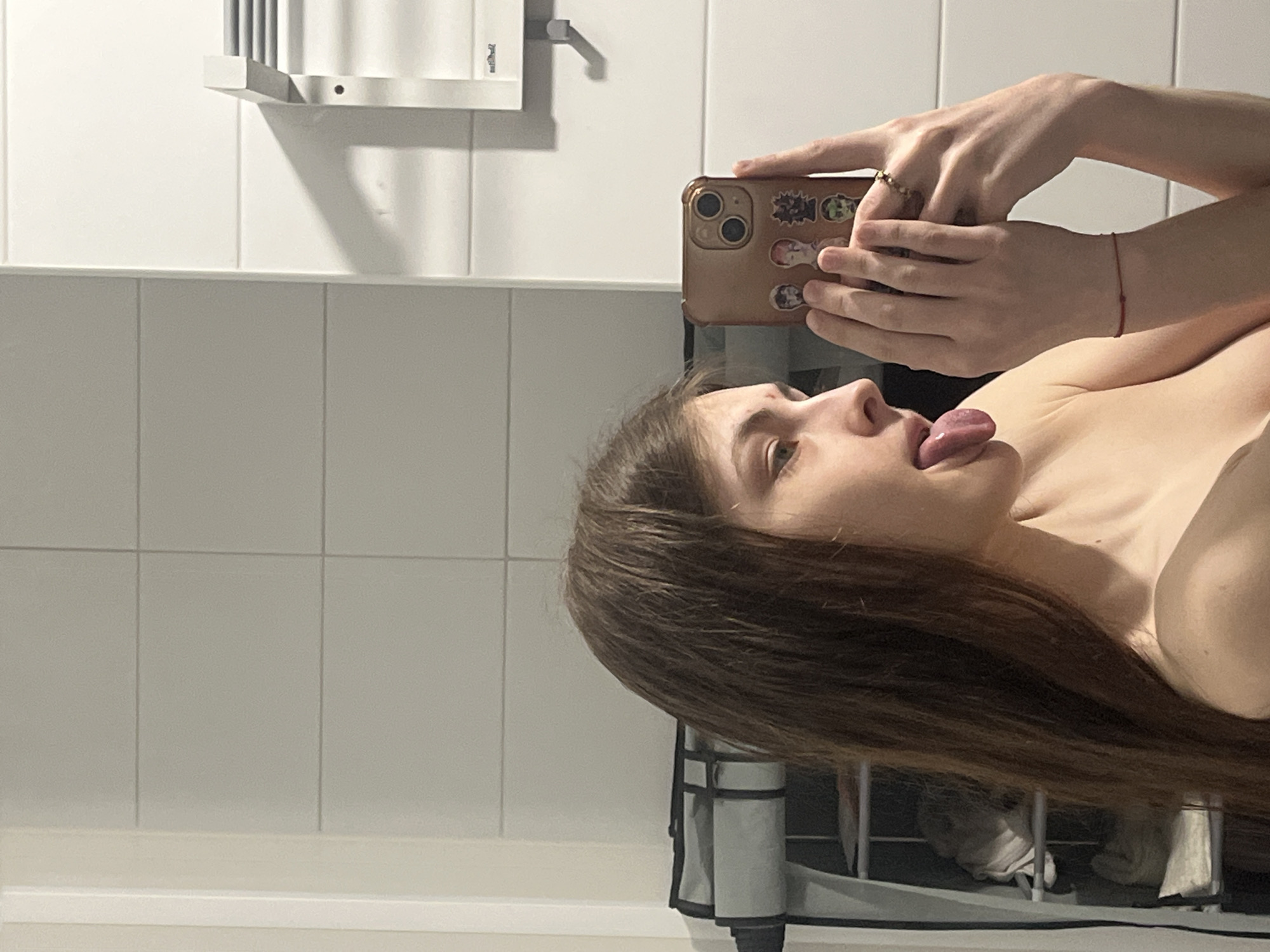 This media file captures a mirror selfie of a young person, likely female, taken in what appears to be a bathroom. The subject is seen in profile, with long brown hair, and is playfully sticking her tongue out. Her expression suggests candidness and humor.
She holds a smartphone with a golden-hued case adorned with several small, colorful stickers, some depicting cartoon or anime characters. A ring is visible on her right hand, and a thin red string bracelet is on her left wrist. The upper part of her bare shoulder/chest is visible.
The background features white and light grey rectangular tiles, common in bathrooms. Above the tiles, a simple metal shelf or rack is mounted. In the lower right, partially obscured, is a dark metal frame, possibly a laundry basket, containing white towels or clothing items. The image is oriented sideways, with the person's head to the left. The scene provides a glimpse into a private, informal moment, set within an indoor environment in Saint Petersburg, Russia. No discernible text is clearly legible on the stickers or in the background.