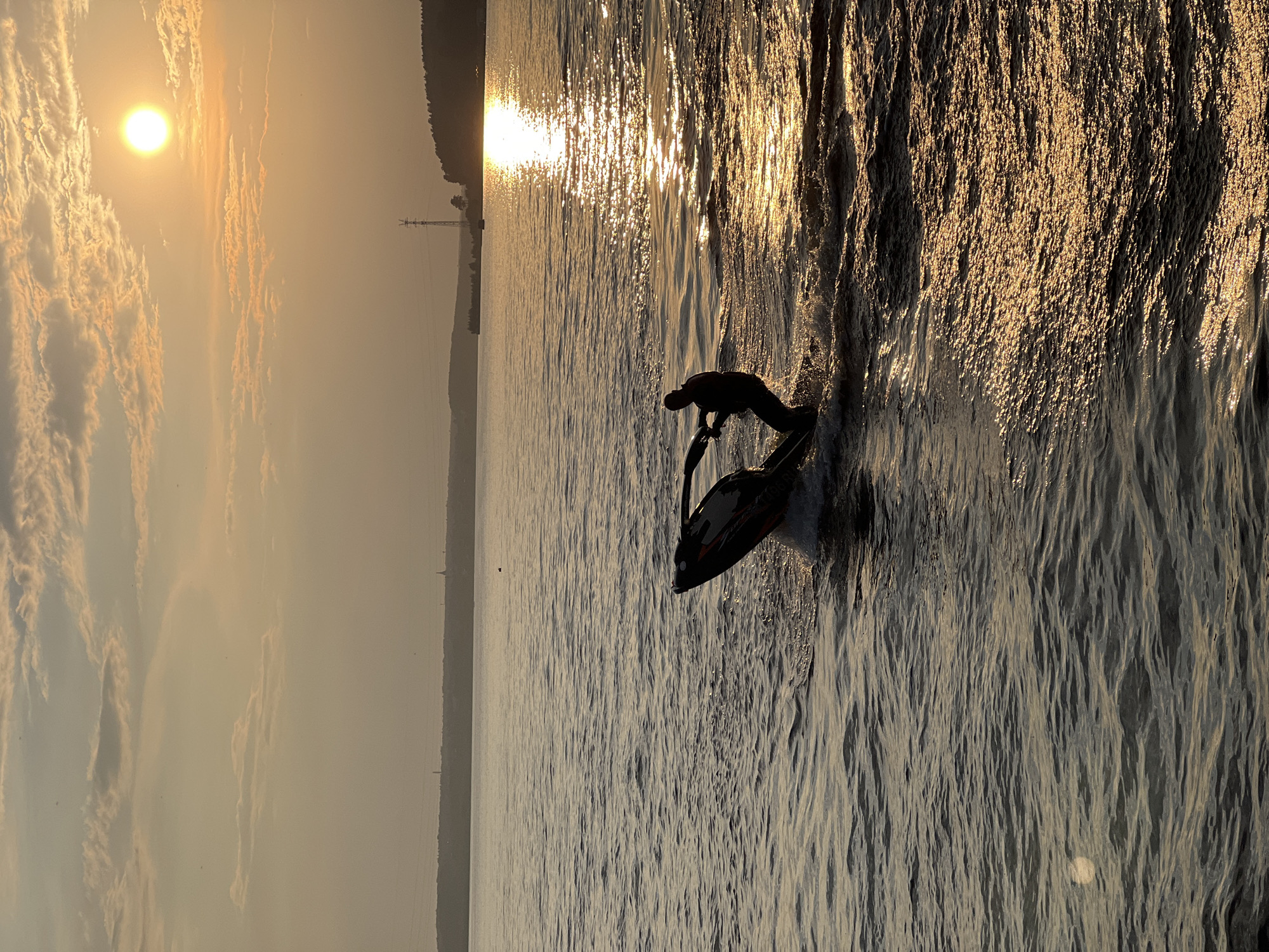 A person rides a jet ski on the Kama River in Izhevsk, Russia, during sunset. The sun is low in the sky, casting a warm golden glow on the water and clouds. The jet ski is dark with orange accents, and the rider is silhouetted against the bright sky. The river is calm, with gentle ripples on the surface. In the distance, the shoreline is visible, with trees and buildings lining the bank. The overall mood of the image is peaceful and serene.