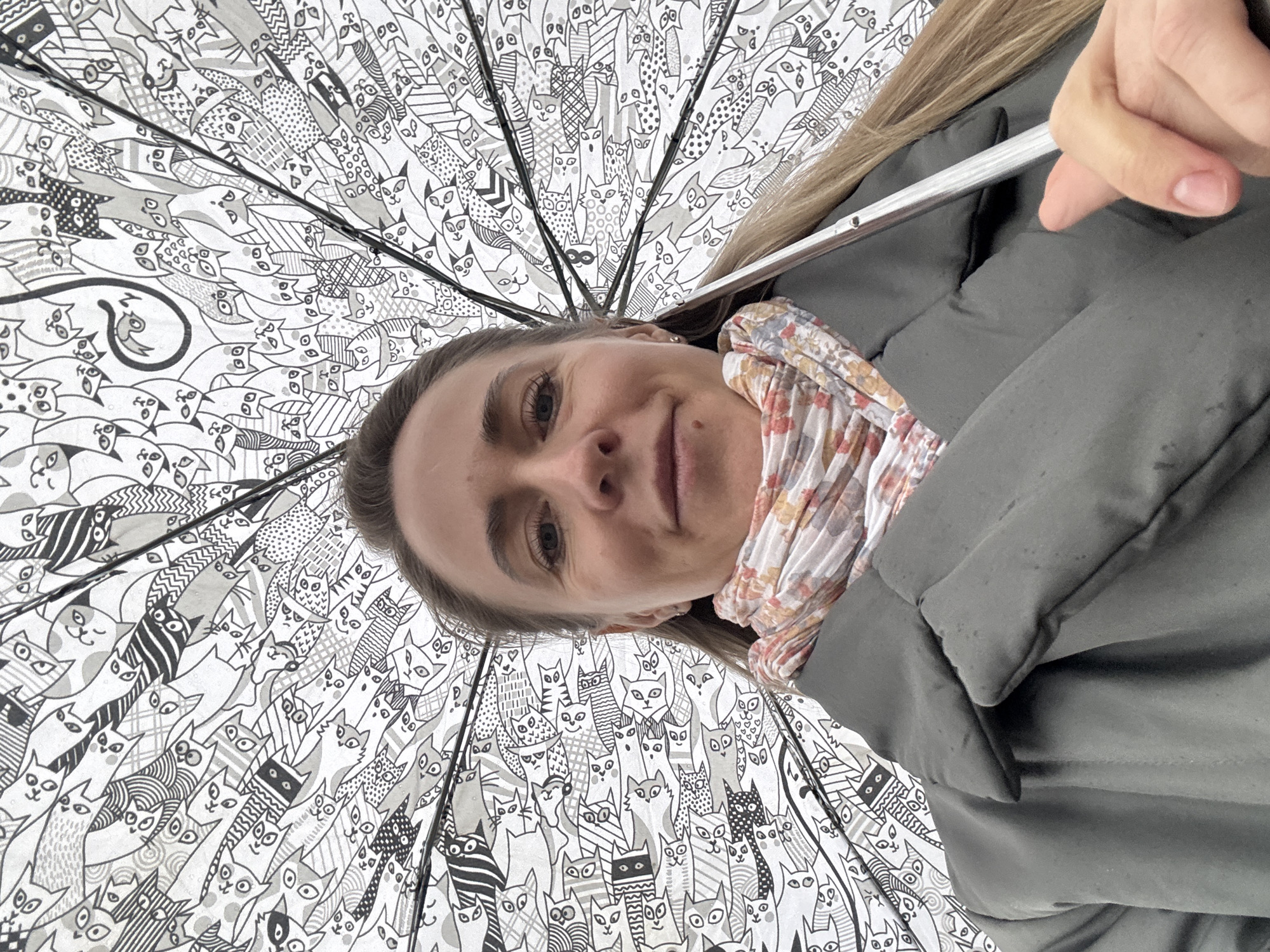 The image shows a woman looking up at the camera from under an umbrella. The umbrella is decorated with a pattern of stylized cats in black and white. The woman has fair skin, light brown hair, and is wearing a patterned scarf and a dark grey jacket. Her hand is holding the umbrella's handle, which is a silver-colored metal rod. The scene's location is not discernible, and there is no way to determine if it is in the city of Chaikovsky, Russia, based solely on the visual information provided.