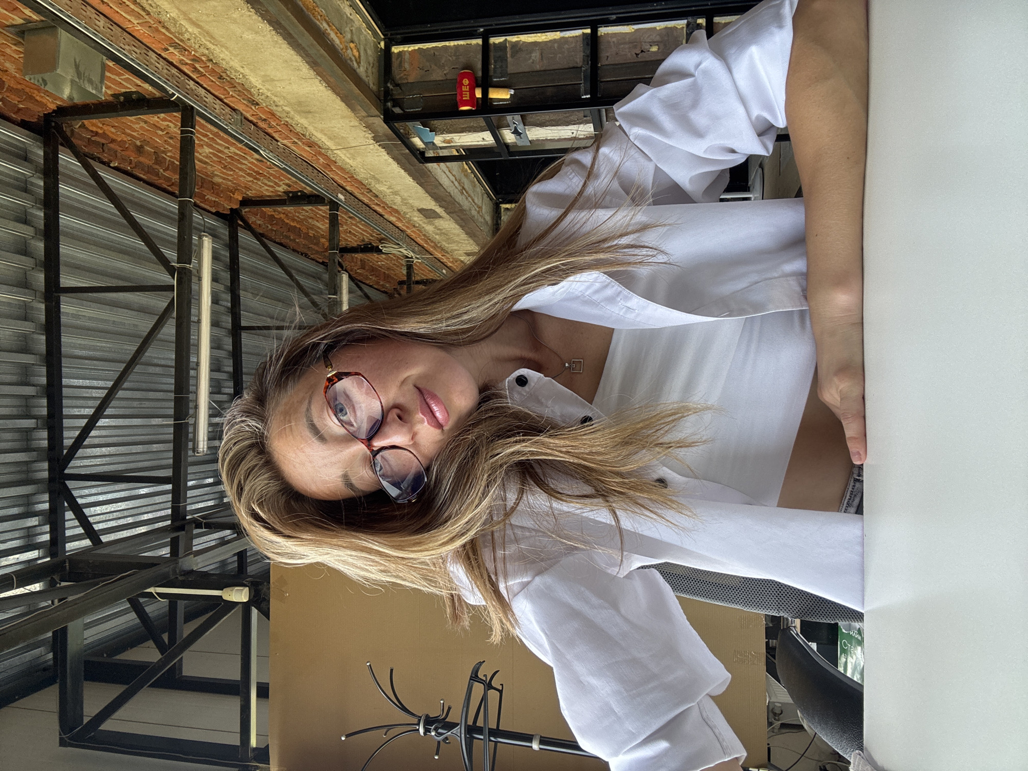 In the city of Chaikovsky, Russia, a woman with long, blonde hair and glasses sits at a white table. She is wearing a white crop top and a white unbuttoned shirt. Her left arm is resting on the table, and her right hand is also on the table. Behind her, a brick wall is visible, along with exposed ceiling beams and fluorescent lights. A coat rack is also present on the left side of the image, and a red object with yellow lettering is mounted on the wall on the right side.