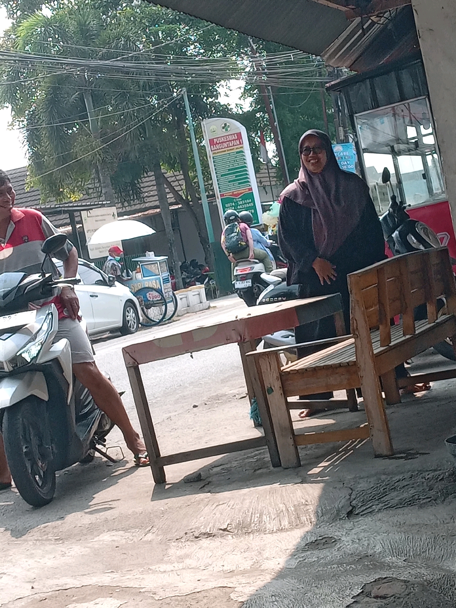The image depicts a street scene in Banguntapan, Indonesia, on a sunny day. In the foreground, a white motorcycle is parked, with a person's legs visible. To the right, a woman wearing a hijab and glasses stands near a wooden table and chairs. Behind her, a white car is parked, and further in the background, there is a street vendor with a cart. Signs and trees are also visible in the environment, suggesting a commercial or residential area. The overall atmosphere is casual and appears to be during daylight hours.