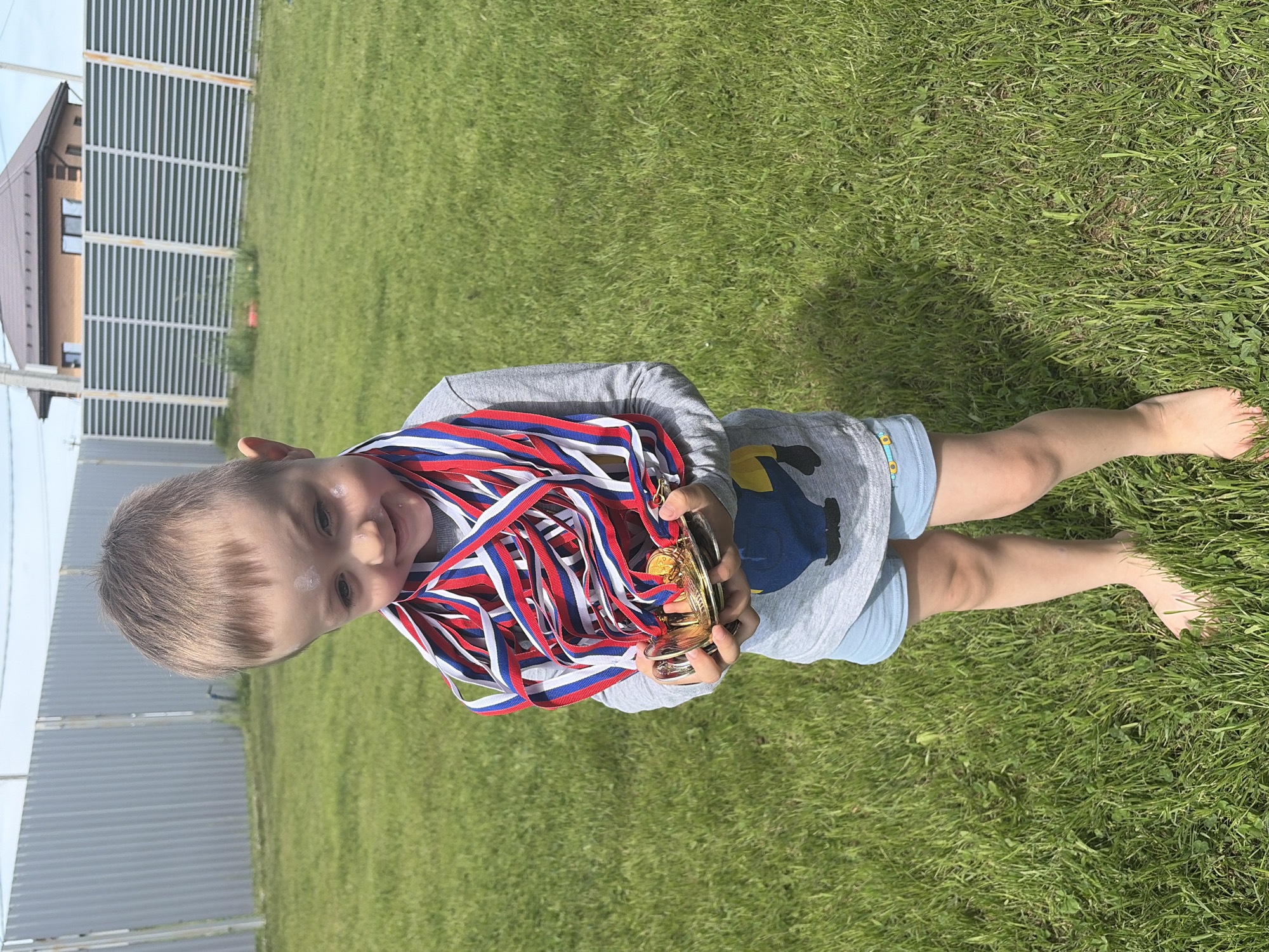 A young child stands barefoot on a lawn, holding a stack of medals and a trophy. The lawn is green. In the background is a fence and a house. The child has blond hair. The child is wearing a gray shirt and light blue shorts. The medals have red, white, and blue ribbons. The location is Chaikovsky, Russia.