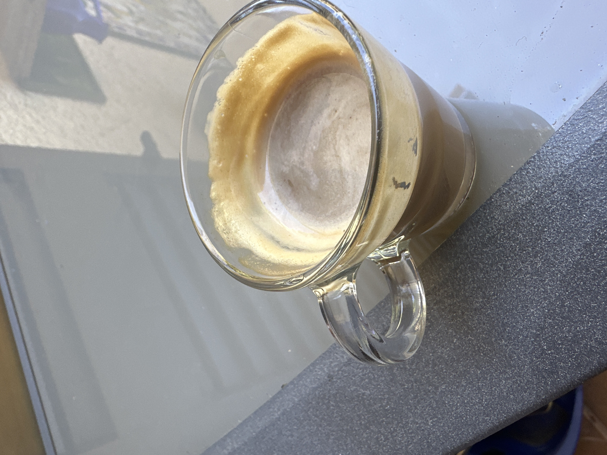 A top-down view shows a partially consumed coffee beverage in a clear glass mug with a handle. The mug rests on a gray, textured surface. To the left of the mug is a light-colored, possibly reflective surface, with shadows visible, suggesting the presence of sunlight.
The photograph appears to have been taken outdoors, possibly on a patio or balcony, but there is insufficient information to confirm the location in the city of Sant Feliu de Guíxols, Spain. There is an absence of individuals, interactions, or specific environmental cues, such as landmarks or geographical features.