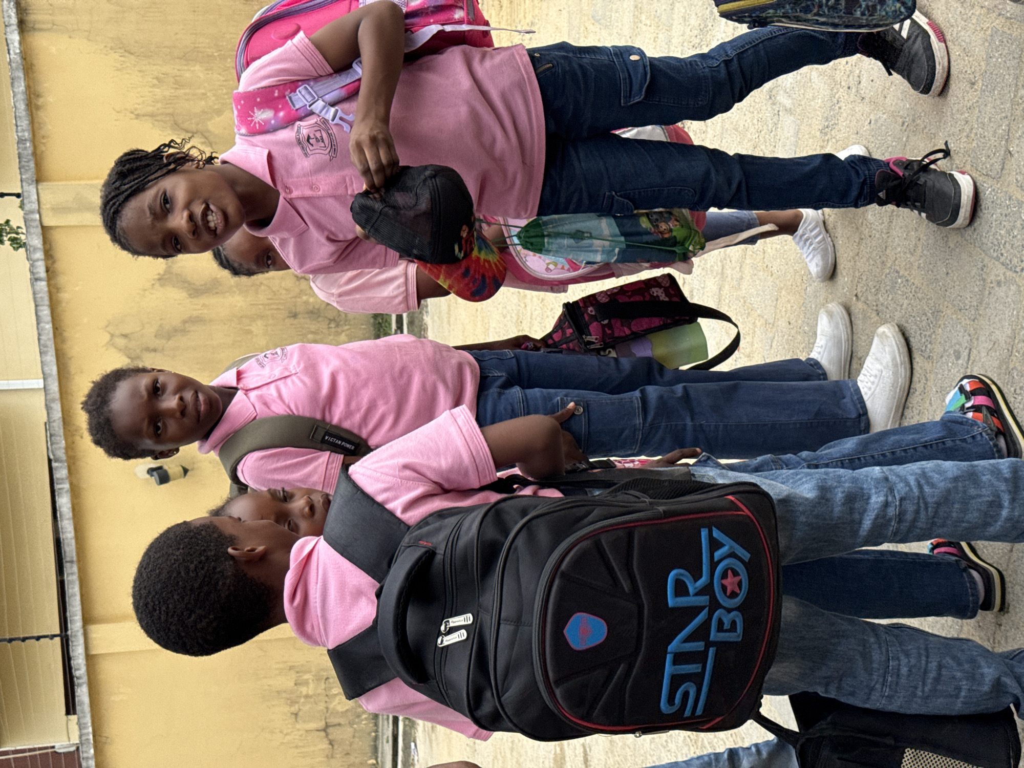 Three children wearing pink shirts and blue jeans stand close together on a paved area. The child in the front wears a black backpack with "STAR BOY" written on it in blue and red letters. The child in the middle wears a backpack with "VICTAN POWER" on the strap. The child in the back holds a black baseball cap and wears a pink backpack with stars on it. A yellow wall is in the background. The scene appears to be outside. This is potentially located in Ado, Nigeria, judging by the prompt.