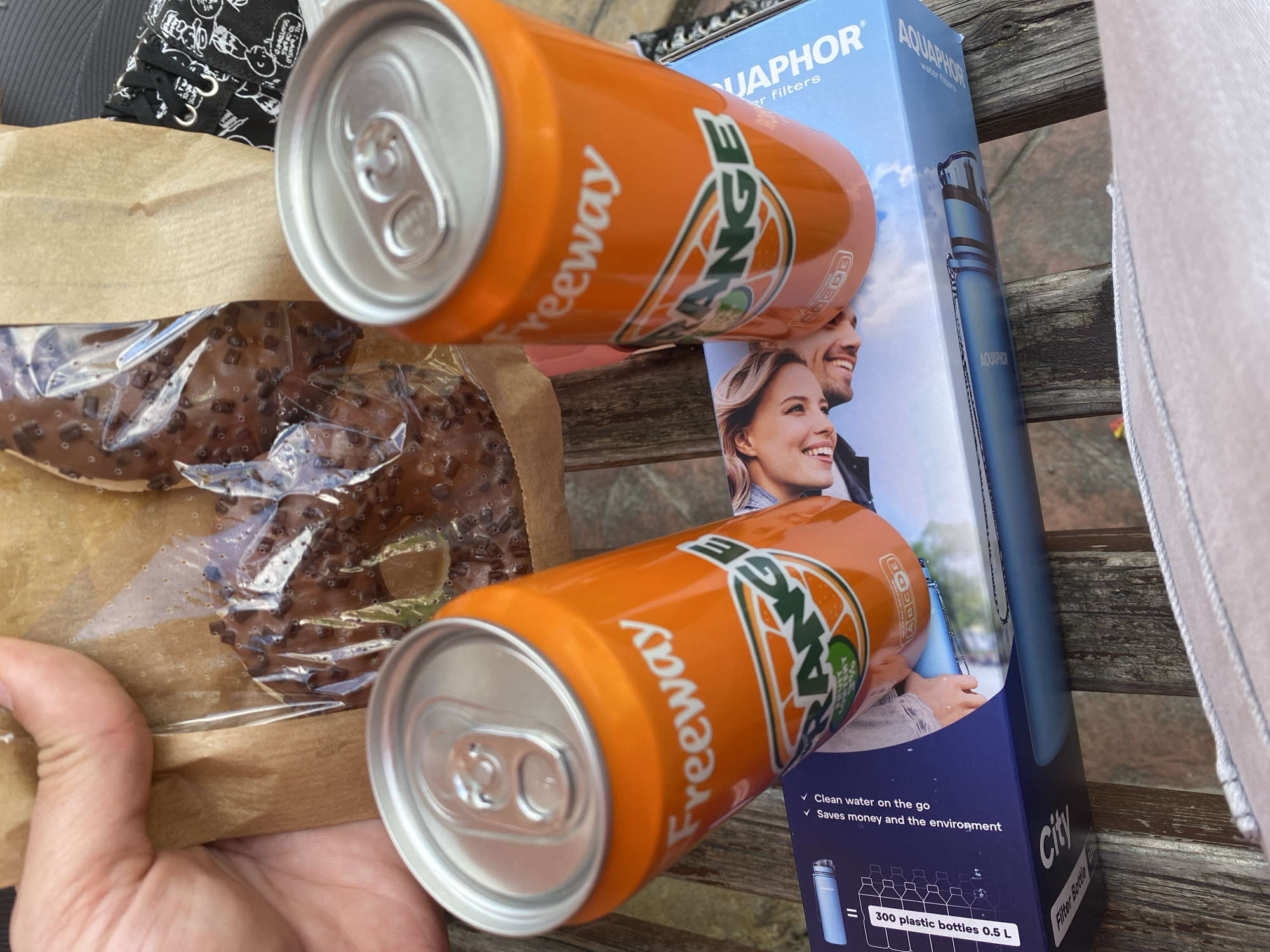 An overhead shot shows a hand holding a brown paper bag with two chocolate-covered pastries inside, resting on a wooden table. Next to the bag are two orange soda cans from the brand "Freeway," with "Orange" printed on them. Below the cans is a blue box that appears to be for a water filter bottle, featuring images of happy people and the brand "AQUAPHOR." The setting is an outdoor cafe or restaurant in the city of Smolyan, Bulgaria, as evidenced by the presence of the unique "City" brand filter bottle on the box. The overall atmosphere suggests a casual outing or a picnic.
