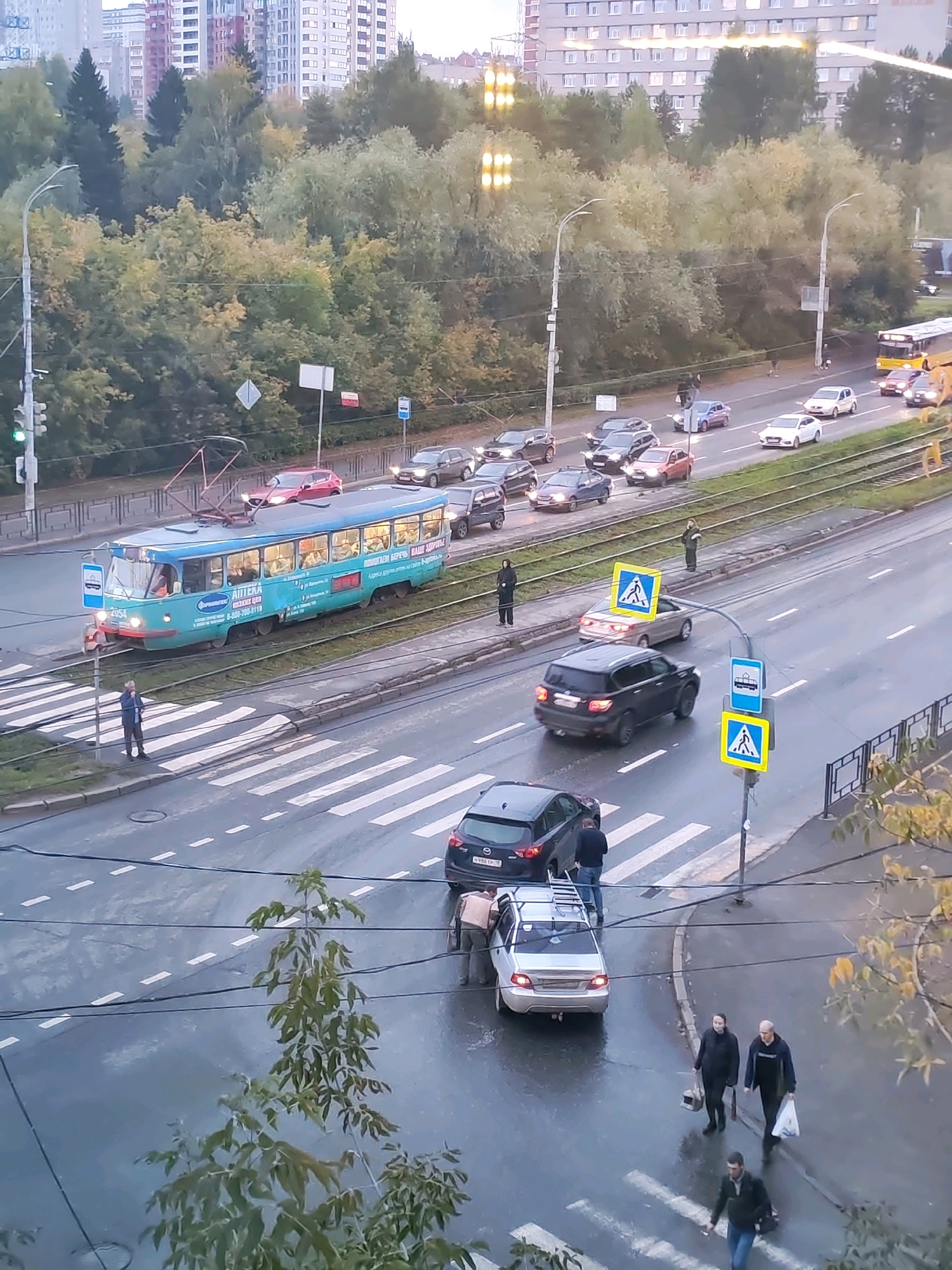 This aerial view captures a street scene in Izhevsk, Russia, under overcast skies, likely during autumn. The road is wet, indicating recent rain. A light blue tram, identified by the number "2054" on its front, is traveling on tram tracks. The tram is decorated with advertisements, including one for "APTEKA."
Several vehicles are present on the road. A silver sedan with a ladder on its roof is stopped at a pedestrian crossing, with three individuals standing nearby, two appearing to be interacting with the car. A dark SUV is also stopped near the pedestrian crossing, and a dark-colored sedan is further ahead. Other cars are visible in the background, forming a traffic flow.
Pedestrians are also visible. One person stands on the sidewalk at a pedestrian crossing, seemingly waiting. Two people are walking across another pedestrian crossing, one carrying a plastic bag. Another individual walks on the sidewalk further down the road. In the background, apartment buildings with many windows line the horizon, and trees with autumn foliage are abundant, framing the scene. Traffic lights are visible, with one showing a green light. The overall impression is a typical urban street in Russia with active traffic and pedestrian movement.