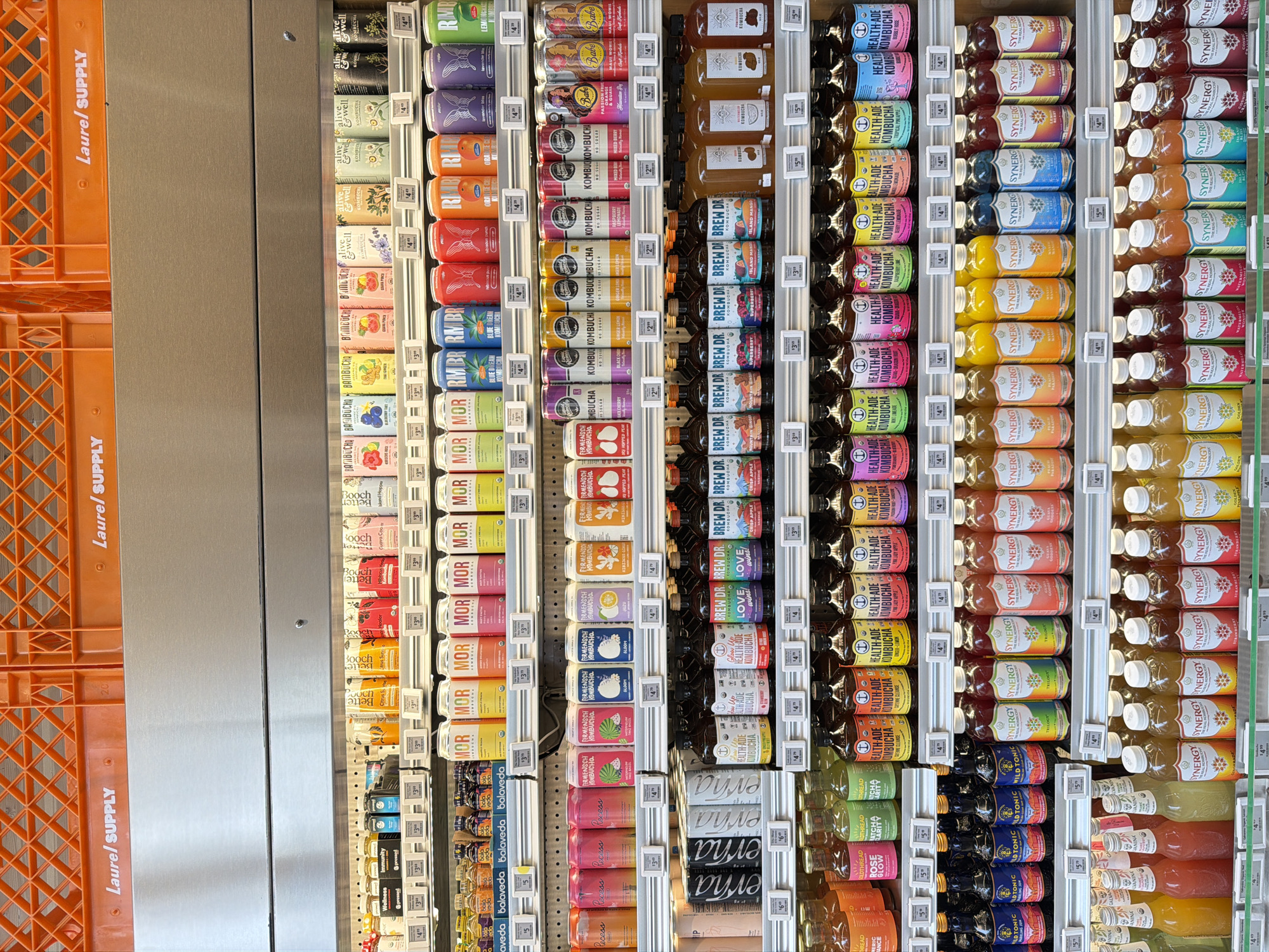 The image shows a refrigerated aisle in a grocery store, likely in West Hollywood, California, United States of America, displaying a variety of beverages. The shelves are stocked with numerous bottles and cans of kombucha and other health drinks. Brands such as "MOR," "REALLY," "BREW DR.," "HEALTH-ADE," "SYNERGY," and "WILD TONIC" are visible. The products are arranged in neat rows, with price tags displayed below each item. The lighting is typical for a retail environment, illuminating the products clearly. The overall impression is one of a well-organized and stocked beverage section of a grocery store.