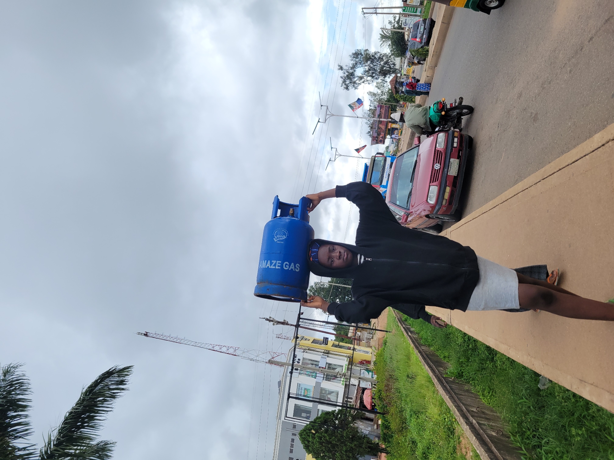 A young person carries a blue gas cylinder labeled "AMAZZE GAS" on their head, walking along a paved walkway. They are wearing a black hooded sweatshirt and a light gray skirt. In the background, there are buildings, a red car, a motorcycle with a rider, and a street with other vehicles. The sky is overcast, suggesting a cloudy day. The scene appears to be on a street in Jos, Nigeria, characterized by its urban setting with pedestrian pathways, roads, and commercial buildings. A communication tower is visible in the distance, and palm fronds overhang from the left side of the image.
