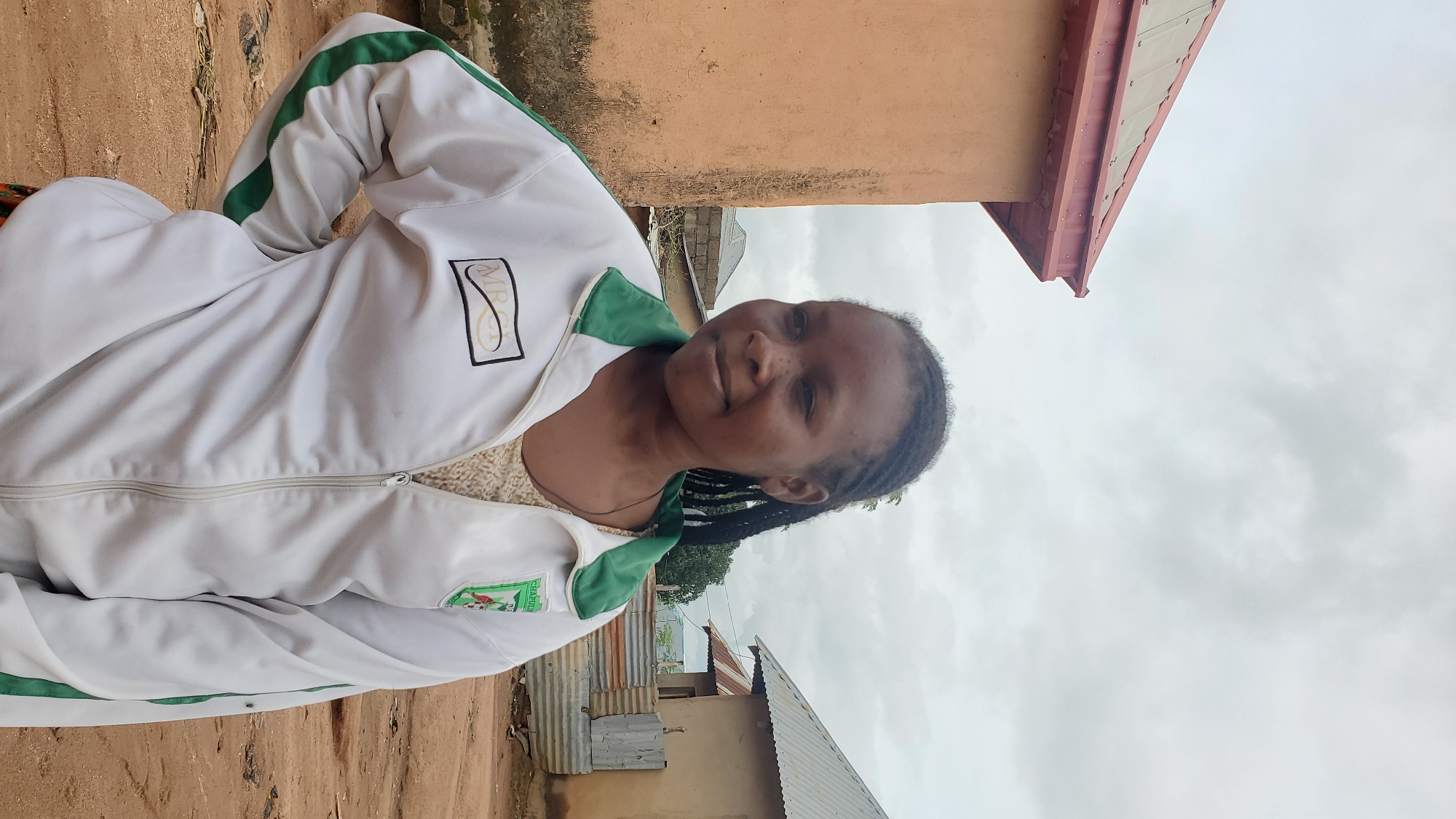 A medium shot captures a young Black woman with braided hair, facing the right side of the frame. She is wearing a white zip-up jacket with green trim and two patches on the chest: one rectangular with black lettering and a logo inside, and another circular patch with green and white details and a soccer ball graphic. A light-colored, textured garment is visible underneath her jacket. Her left hand is placed on her hip, and her right arm hangs loosely. The background shows a rural or semi-urban setting in Jalingo, Nigeria. To the left, a portion of a building with a reddish-brown, metal-clad roof and an orange-colored wall is visible. To the right, another building with a similar metal roof and light-colored walls stands, with corrugated metal sheeting and wooden planks creating a partition in front of it. The ground is unpaved, with scattered debris. The sky is overcast with gray clouds.