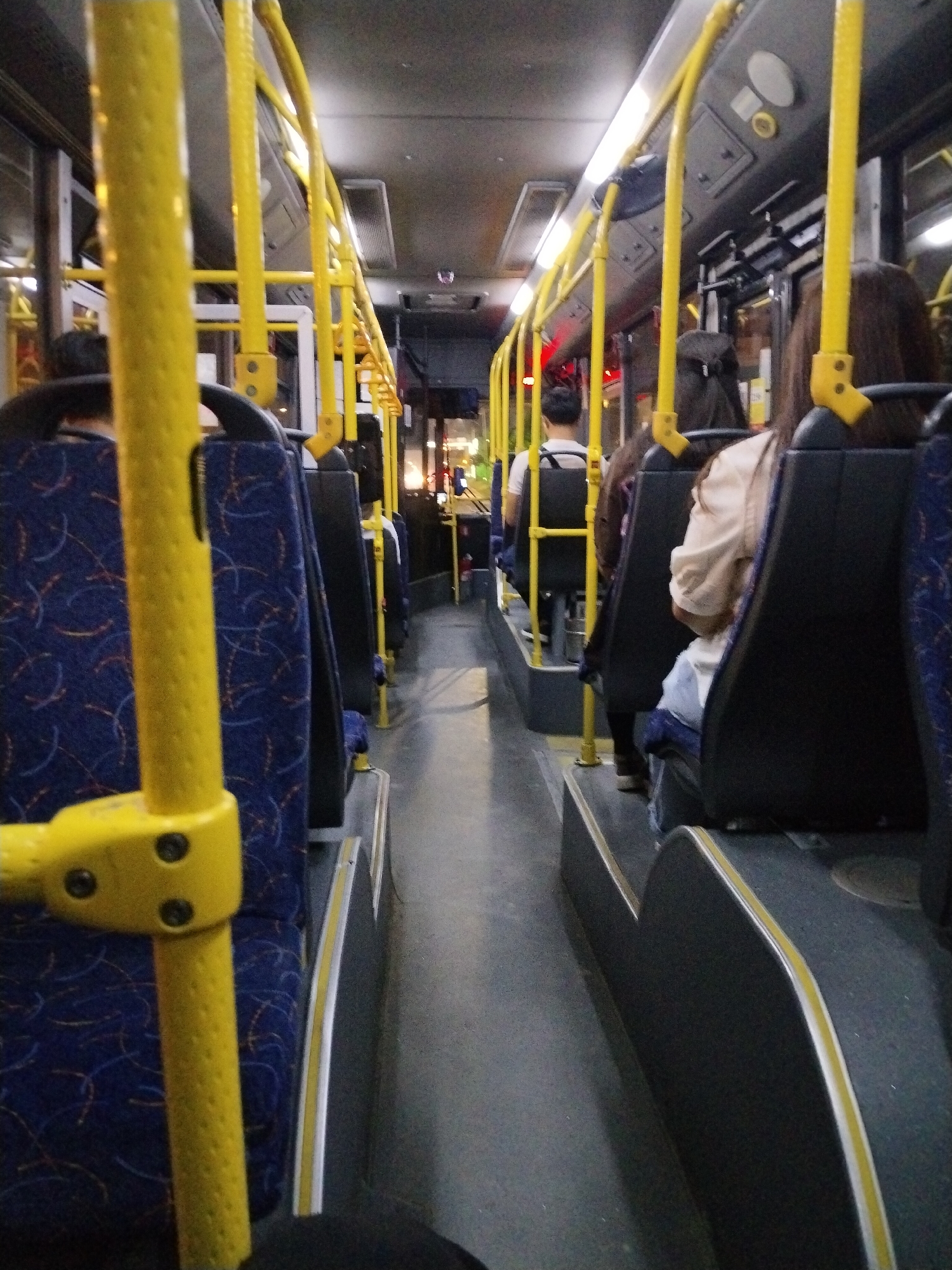 The image shows the interior of a public bus. Yellow poles and handrails are prominent throughout the bus. Several passengers are seated in blue and black patterned seats. The lighting is dim, with overhead lights visible. The flooring appears to be dark gray. The scene suggests a typical commute on public transportation.