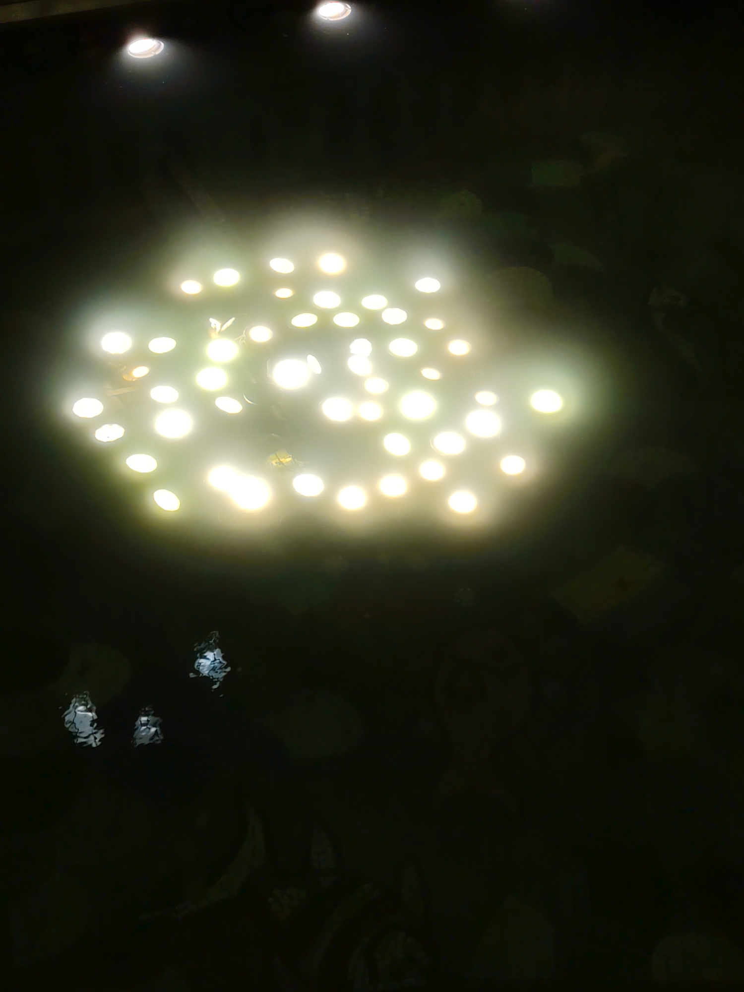 In an outdoor night setting, a cluster of bright spotlights illuminate a dark surface, likely water. The lights are concentrated in the upper center of the image, creating a hazy glow that obscures details beneath. In the lower portion of the image, three small, indistinct reflections appear on the dark surface, suggesting the presence of other light sources or objects. The overall scene is dark and mysterious, with the prominent spotlights dominating the visual field. The location is Unknown, Unknown.