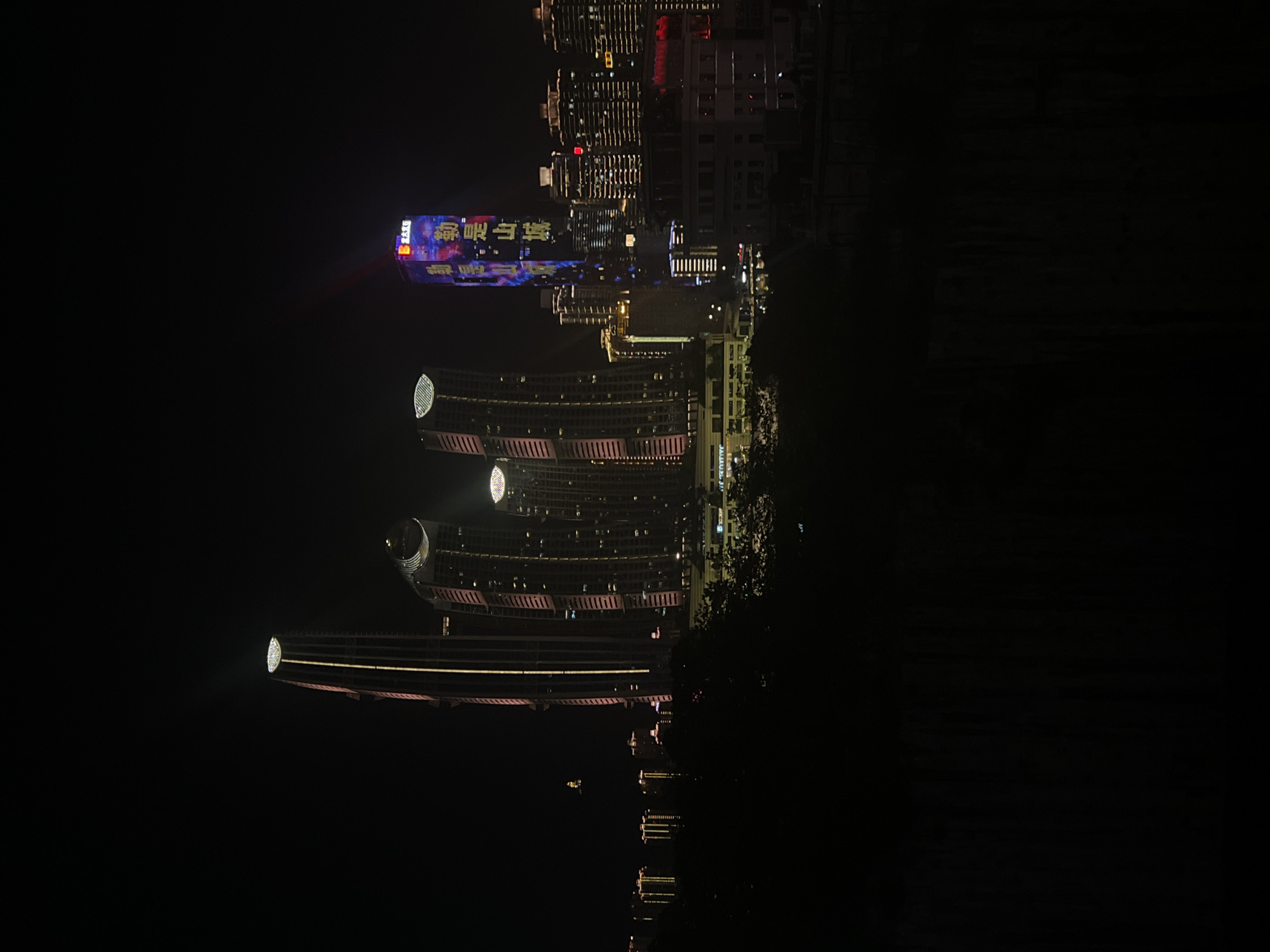 This media file displays a nighttime view of a cityscape, with buildings illuminated by colorful lights. The image appears to be taken from a slightly elevated perspective, looking out over a body of water that reflects the lights of the buildings. The overall scene is one of urban beauty and nighttime activity.
The main subjects are the modern architecture of several large buildings. One prominent building features a large digital display with illuminated text in what appears to be Chinese characters. The text is vibrant and colorful, creating a focal point in the image. Other buildings showcase intricate lighting designs along their facades, highlighting their architectural features. The reflections in the water below add a sense of depth and symmetry to the scene.
The time of day is clearly night, as indicated by the darkness of the sky and the artificial illumination of the city. There are no visible signs of weather conditions; the sky is dark and appears clear. The presence of illuminated buildings and the overall ambiance suggest a bustling urban environment. The location context, Jiangbeicheng Street in China, further supports the interpretation of a modern Chinese city.
No people are directly visible in the image, suggesting a focus on the architectural landscape and the city's nighttime illumination rather than human presence. The mood conveyed by the image is one of wonder and perhaps a sense of awe at the scale and beauty of the illuminated cityscape. The vibrant colors and the reflections create a visually striking and somewhat magical atmosphere.