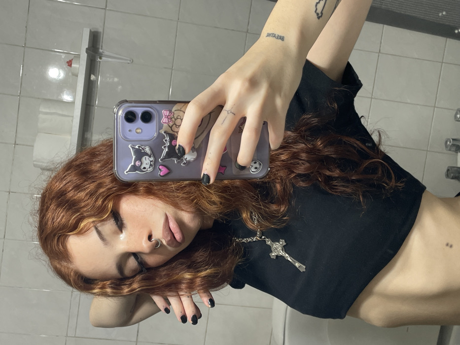 A mirror selfie depicts a young individual, appearing female, in a bathroom located in Buenos Aires, Argentina. The subject has long, wavy, reddish-brown hair with lighter blonde streaks and is wearing a black cropped t-shirt. A silver-colored chain necklace with a crucifix pendant is visible around the neck. The individual features a septum piercing and dark, elongated eyelashes. Black nail polish is applied to the fingernails. Tattoos include a faint horizontal line on the left middle finger, indistinct text on the left wrist, and an abstract design on the left forearm. The subject is holding a light purple iPhone, equipped with a clear case adorned with Kuromi character stickers, in the left hand to capture the reflection. The right arm is bent with the hand resting near the head. The individual is looking down towards the phone with slightly parted lips. The background, reflected in the mirror, shows a wall composed of light-colored square tiles with dark grout. A white shelf is mounted on the wall, holding several rolls of toilet paper and a small red and white object. Partially visible fixtures include the edge of a white porcelain sink and two chrome faucet handles at the bottom of the frame. A dark grey textured object, possibly a bathmat, is also partially visible in the reflection on the lower right.