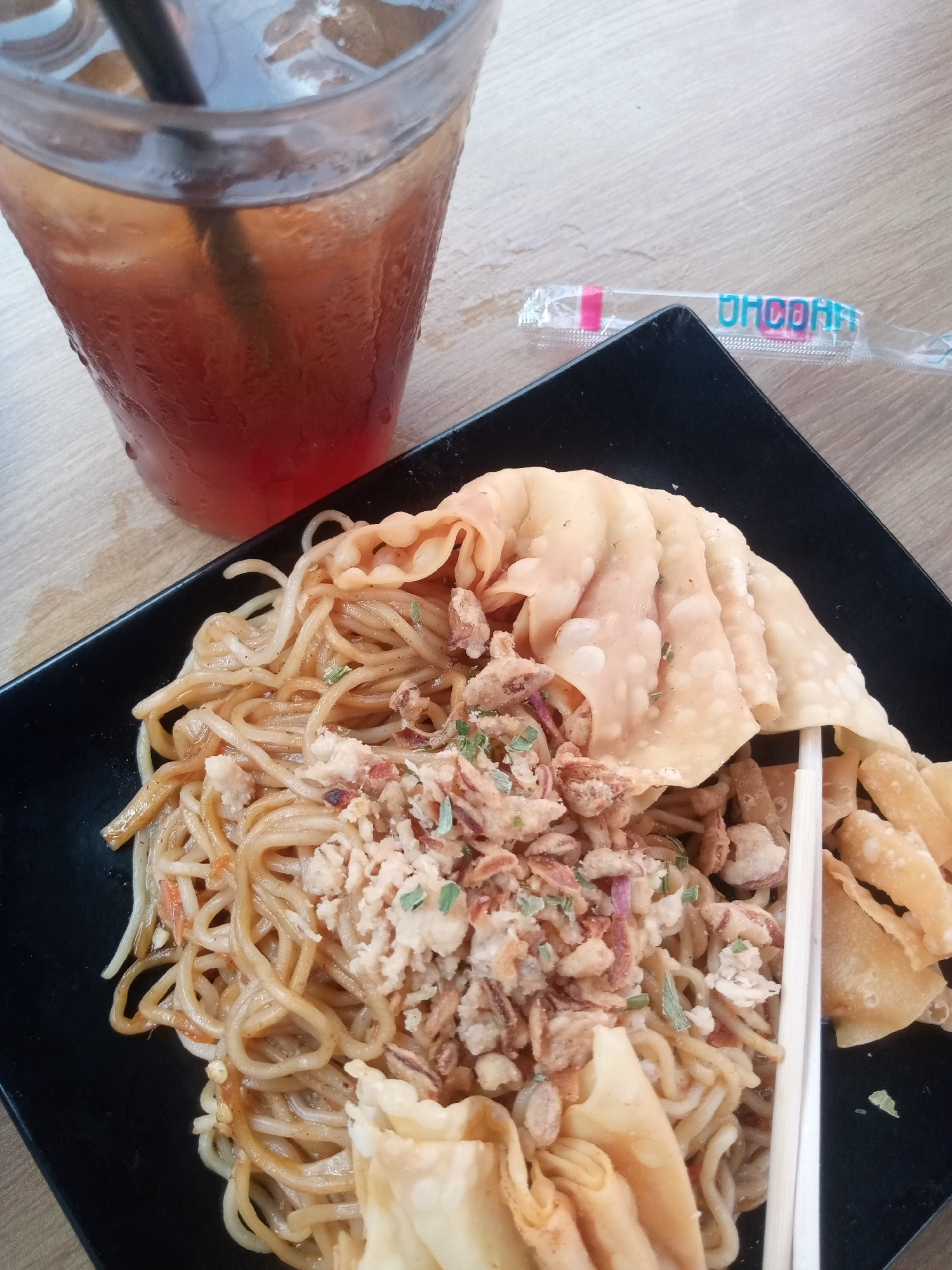 A close-up shot shows a dish of noodles and fried wontons served on a black plate, placed on a wooden table. Beside the plate is a glass of iced tea with a black straw. A clear plastic wrapper with colorful lettering, reading "UHCONH", is also visible on the table, near the edge of the plate. The scene appears to be a meal at a restaurant or food stall in Sukaraja, Indonesia, given the presence of common Indonesian dishes and the packaging of the wrapper. The lighting suggests it is daytime, possibly indoors with natural light.