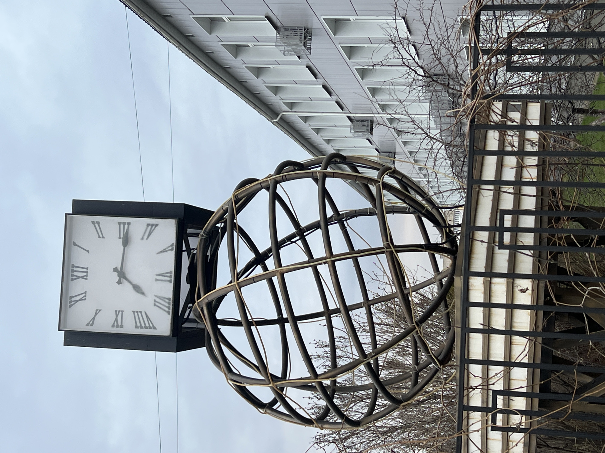 The image shows a view in Chaikovsky, Russia, featuring a clock and metal globe structure outdoors. The clock face is square with Roman numerals, displaying a time around 4:38. Adjacent to the clock is a large, spherical structure made of curved metal rods, resembling a cage or a stylized globe. In the background, there is a building with multiple white-framed windows. A black metal fence is also visible, partially obscured by bare tree branches. The sky is overcast. The picture is taken from a slightly low angle.