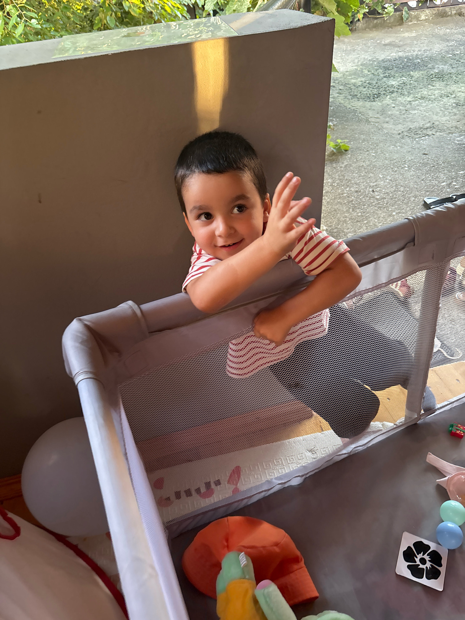 A young boy with dark hair and brown eyes is leaning over a grey playpen, his right arm extended and his hand raised as if waving. He is wearing a red and white striped t-shirt. The playpen has a mesh fabric on its sides and a padded grey frame. Inside the playpen, a dark shadow is visible, possibly a person or a large toy. On the grey surface outside the playpen, a white balloon, an orange hat, a stuffed toy, and other children's items such as a small toy gift box and pastel-colored eggs are scattered. In the background, there is a grey wall casting a shadow, and beyond that, greenery from plants and a glimpse of a paved outdoor area. The lighting suggests it is daytime.