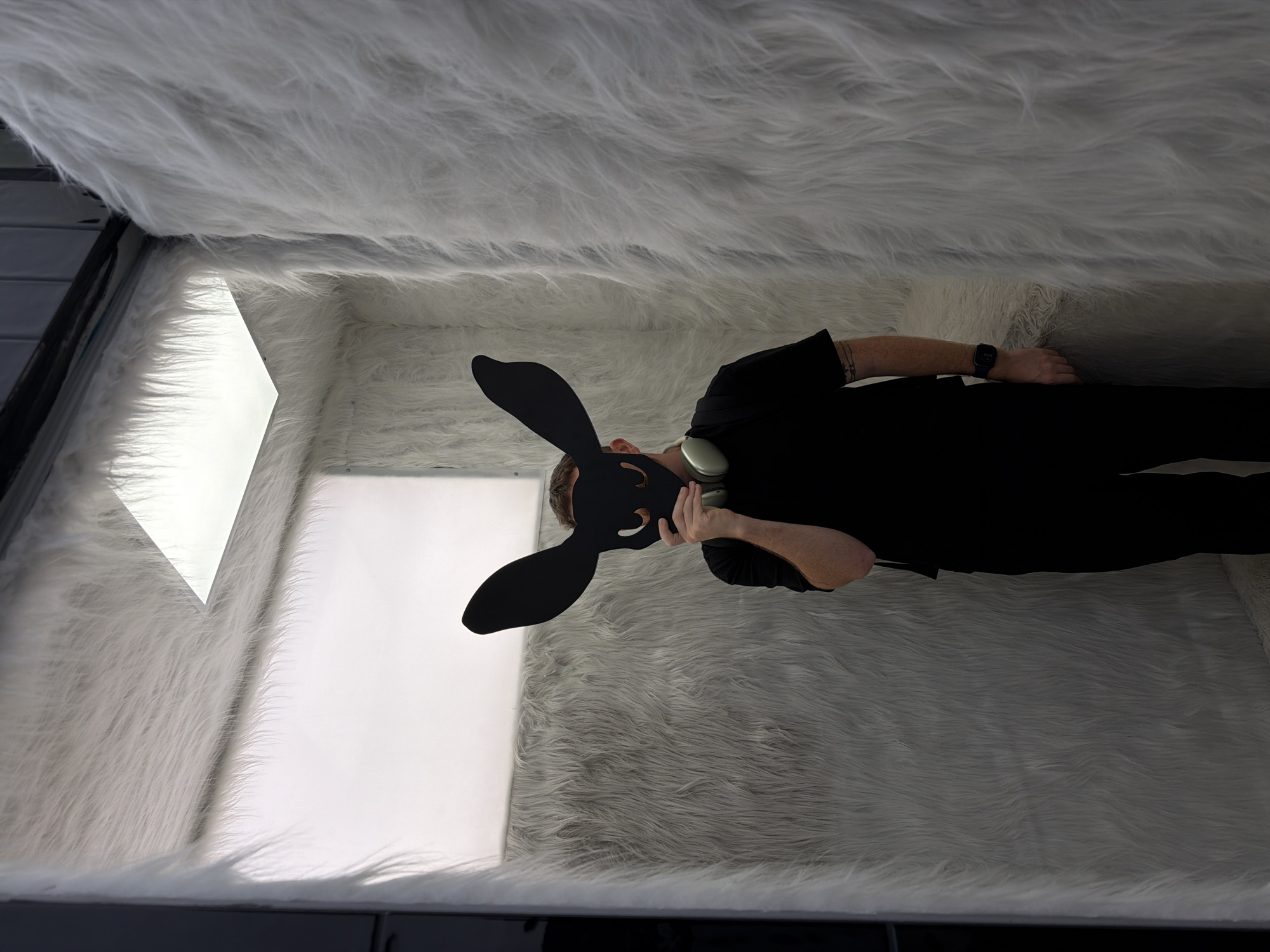 A person wearing a black rabbit mask and dressed in black is lying down in a bright, enclosed space lined with white faux fur. The person appears to be holding a round, silver object near their face. A wristwatch is visible on their left wrist. The location is not identifiable in the image.