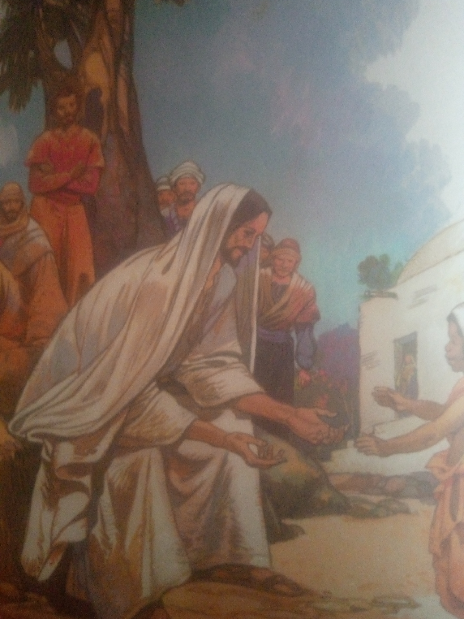 The image depicts a biblical scene, likely the feeding of the multitude, rendered in a painted style. The central figure, dressed in a white robe and head covering, appears to be Jesus. He is kneeling and holding out his hands, seemingly distributing food to a group of people gathered around him. To the left, a man in a red tunic stands beneath a large tree. Other individuals, some with head coverings, are present in the background, observing the event. In the middle ground, a woman in a peach-colored garment is receiving something from Jesus. To the right, a building with a doorway is visible. The sky is a soft blue with hints of clouds. The overall atmosphere appears serene and communal. The scene's setting suggests an outdoor gathering, possibly in a village or rural area, with elements like the tree and the building providing context. The time of day is indeterminate, but the lighting suggests daylight. No text is visible in the image.