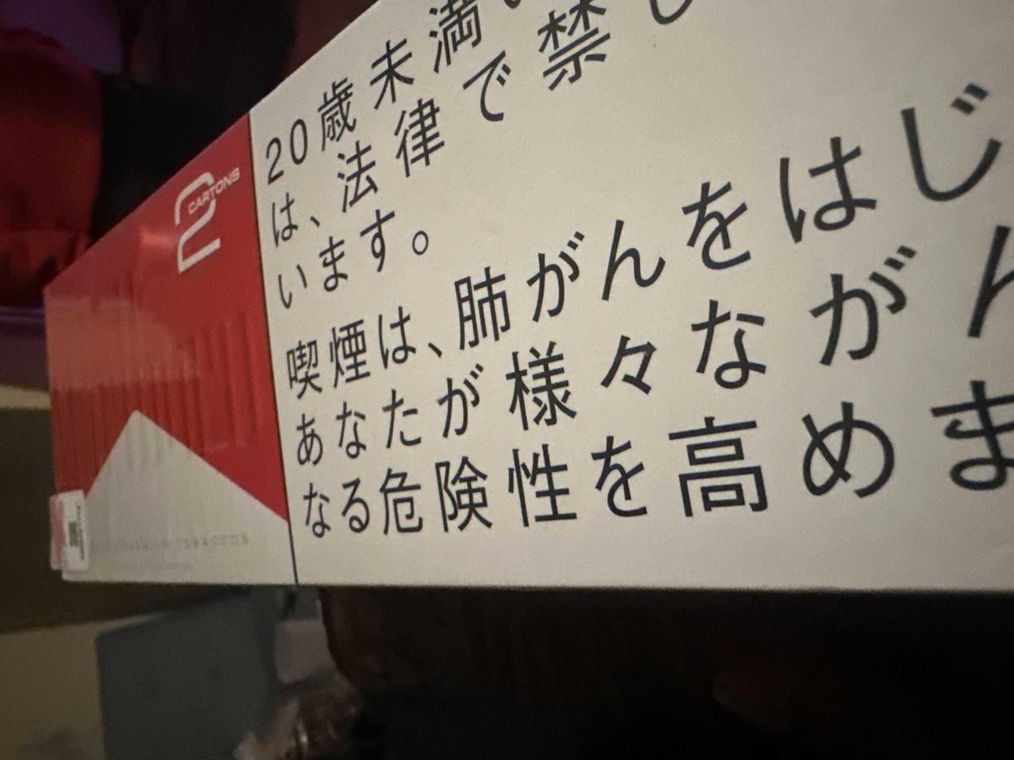 The image shows a white and red cigarette carton from the brand "2 CARTONS". The carton has Japanese writing on its side with the message, "Smoking causes lung cancer."