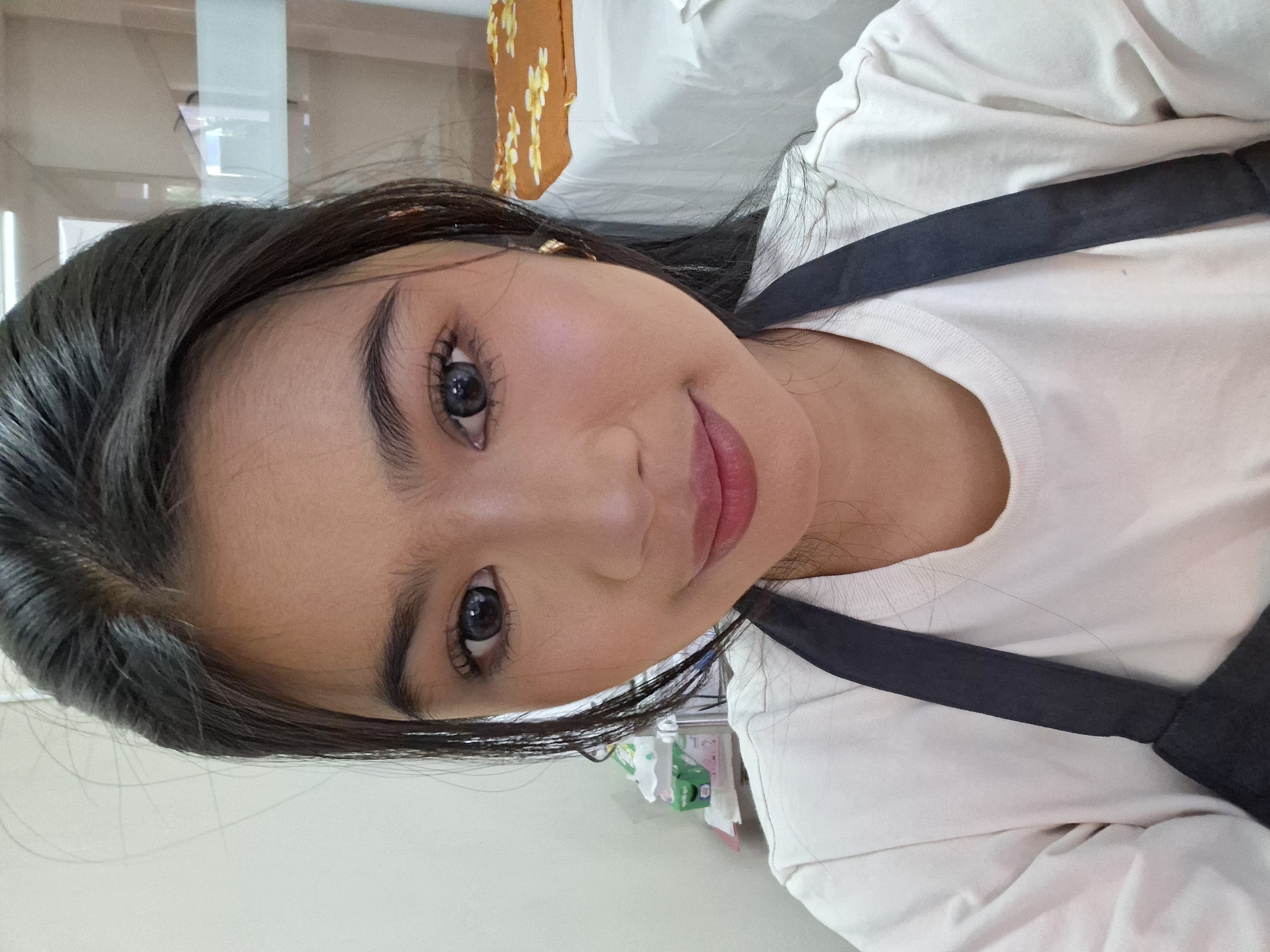 A young woman with dark hair, wearing a white shirt and a black apron, smiles at the camera. She has makeup on, including mascara, blush, and lipstick. Her hair is pulled back, with some strands framing her face. The background is out of focus, but appears to be an indoor setting, possibly a spa or salon, with a white bed and a shelf with tissue boxes on the left. The lighting is bright and even, suggesting it might be daytime. The image is a close-up selfie taken from the chest up. The location in Kuta, Indonesia is not discernible from the image itself.