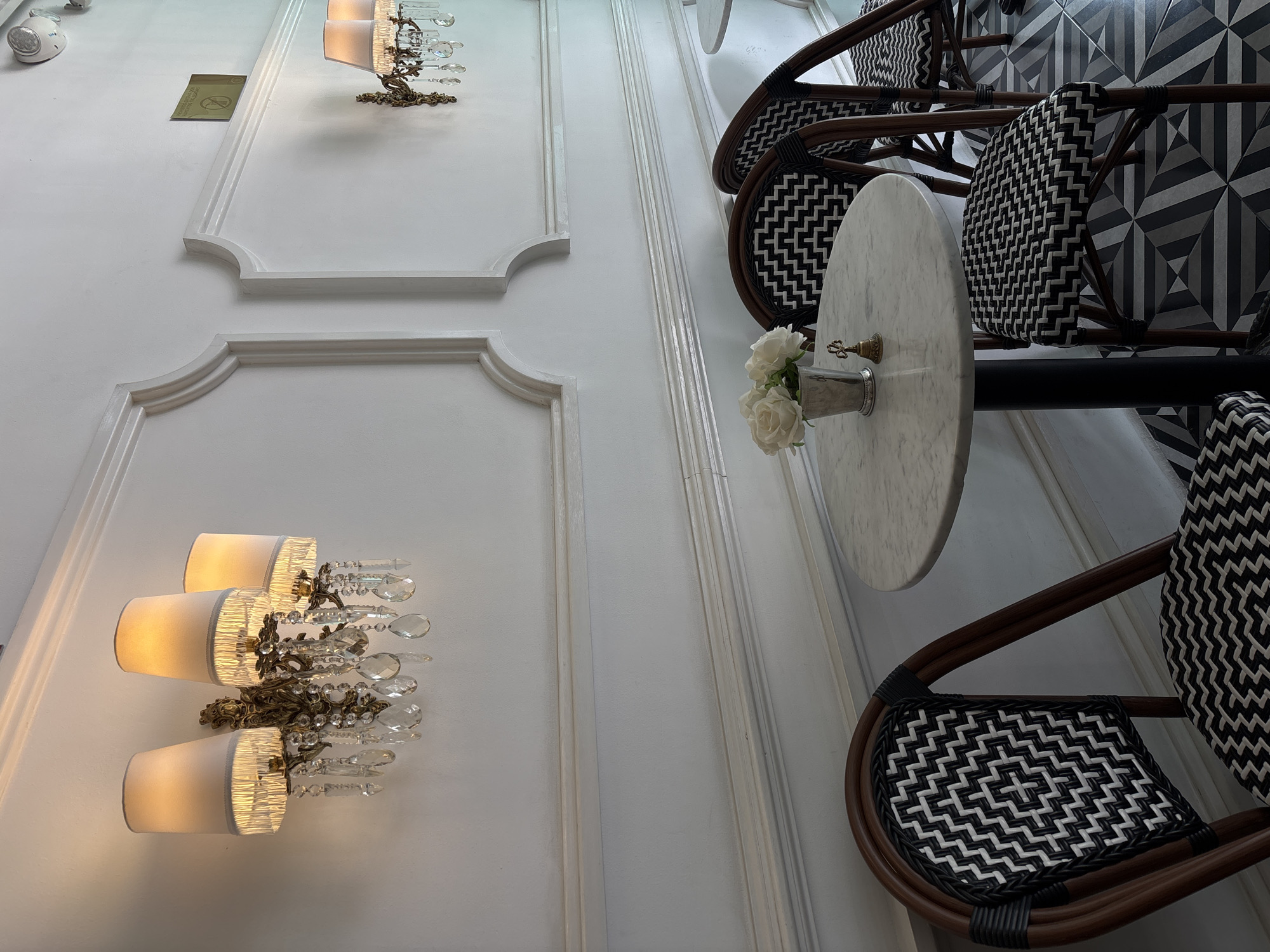A low-angle shot depicts a section of an interior, likely a cafe or restaurant, in San Bernardino Tlaxcalancingo, Mexico. The left side of the frame is dominated by a white wall with decorative molding. Two ornate light fixtures, featuring crystal embellishments and fabric shades, are mounted on the wall.
The right side of the image features a small, round marble-topped table with a vase of white flowers on it. Around the table are woven bistro chairs with dark wood frames and black and white patterned seats and backrests. The floor is tiled with a geometric black and white pattern.