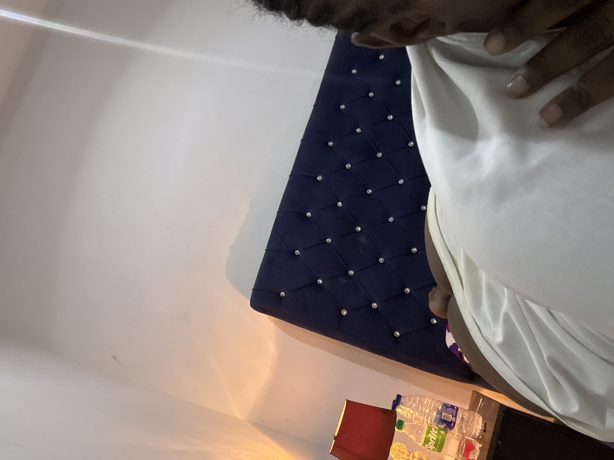 The image shows an individual, possibly male, wearing a white shirt. The person is next to a dark blue, diamond-patterned, upholstered headboard with embedded rhinestones. To the side are several plastic water bottles and a bottle of Sprite on a table. A red lamp is also visible. The location is likely a room in a house or hotel, but there is no evidence to indicate that the location is Abuja, Nigeria.