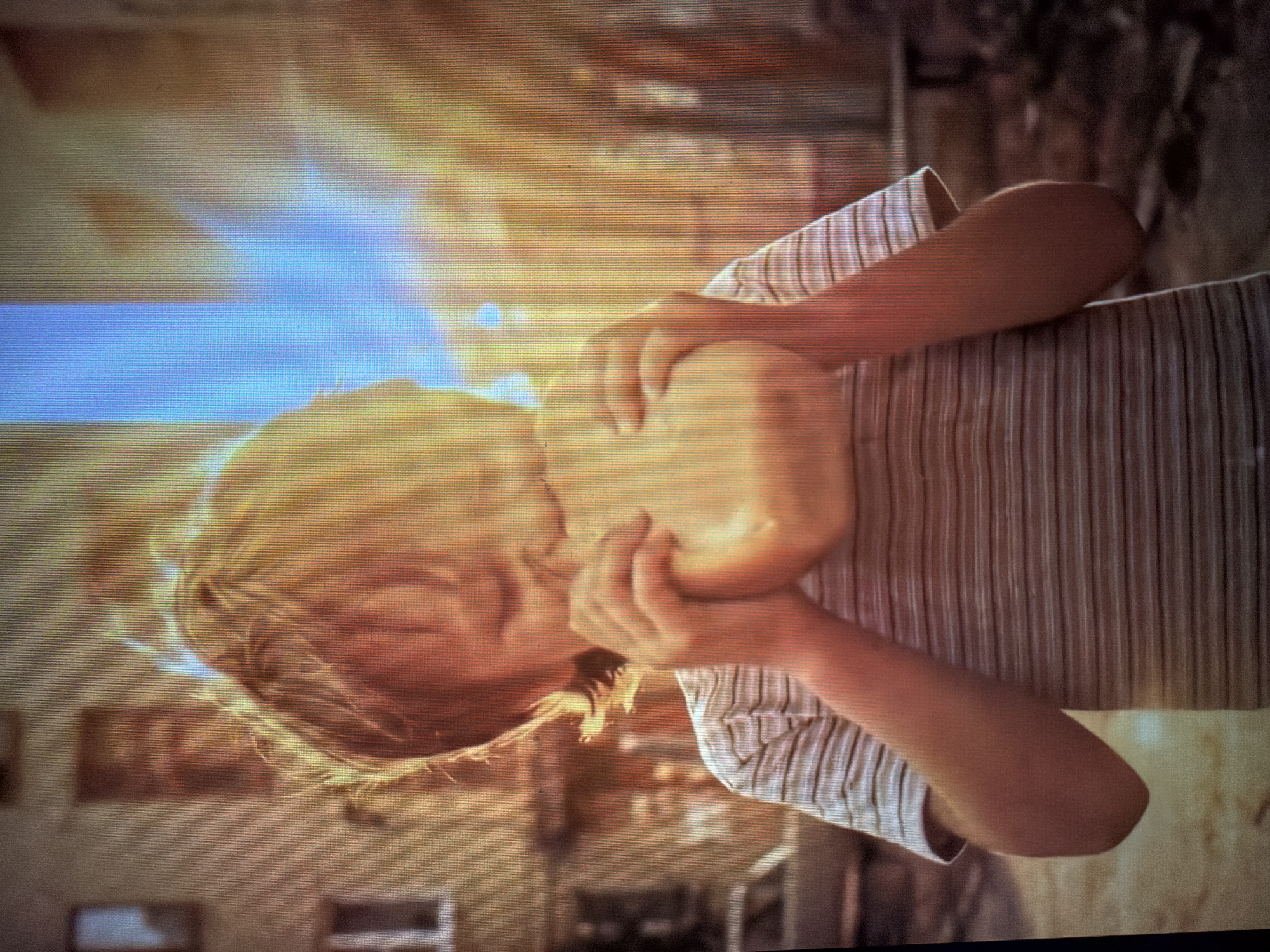 A young child with blond hair is eating a large loaf of bread outdoors in an urban environment. The child is wearing a striped t-shirt and their face is partially obscured by the bread as they take a bite. Sunlight is filtering through the background, creating a bright, hazy effect. Buildings and a parked car are visible in the background, suggesting a city street or courtyard. The overall impression is one of a candid moment captured in natural light.
