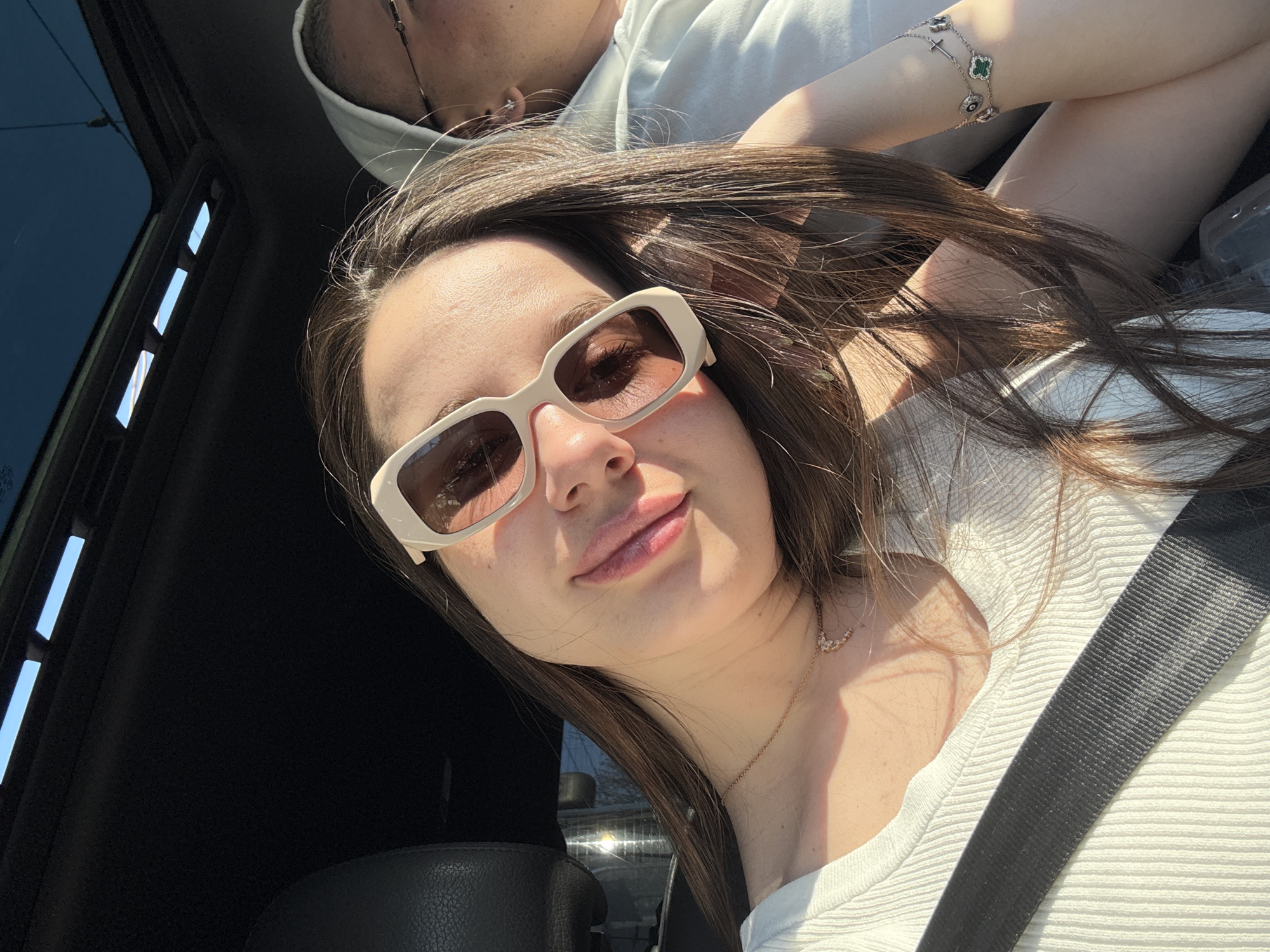 A woman is in a car. The image is a selfie of the woman wearing sunglasses and a white shirt with a seatbelt across her chest. She is also wearing a necklace and a bracelet. Another person wearing a white hoodie is in the background. The location is unclear, so a city cannot be confirmed.