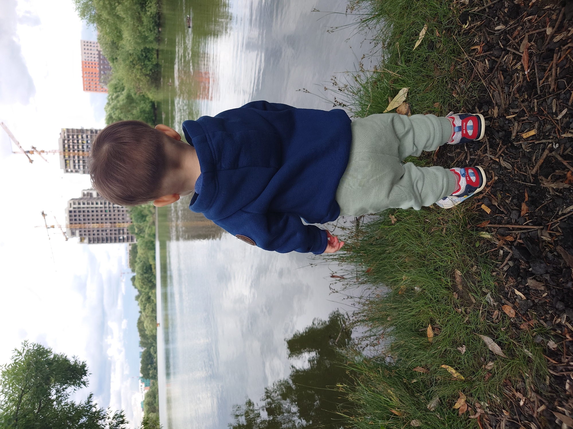A young child, seen from behind, stands on the grassy bank of a body of water. The child is wearing a blue hooded jacket and light green pants. Their shoes are visible, featuring blue, red, and white colors. The water reflects the overcast sky and some of the surrounding trees. In the background, construction cranes and unfinished buildings are visible, indicating an urban or suburban environment. The scene is located in the city of Kommunarka, Russia, characterized by the presence of both natural elements like water and vegetation, and ongoing urban development.