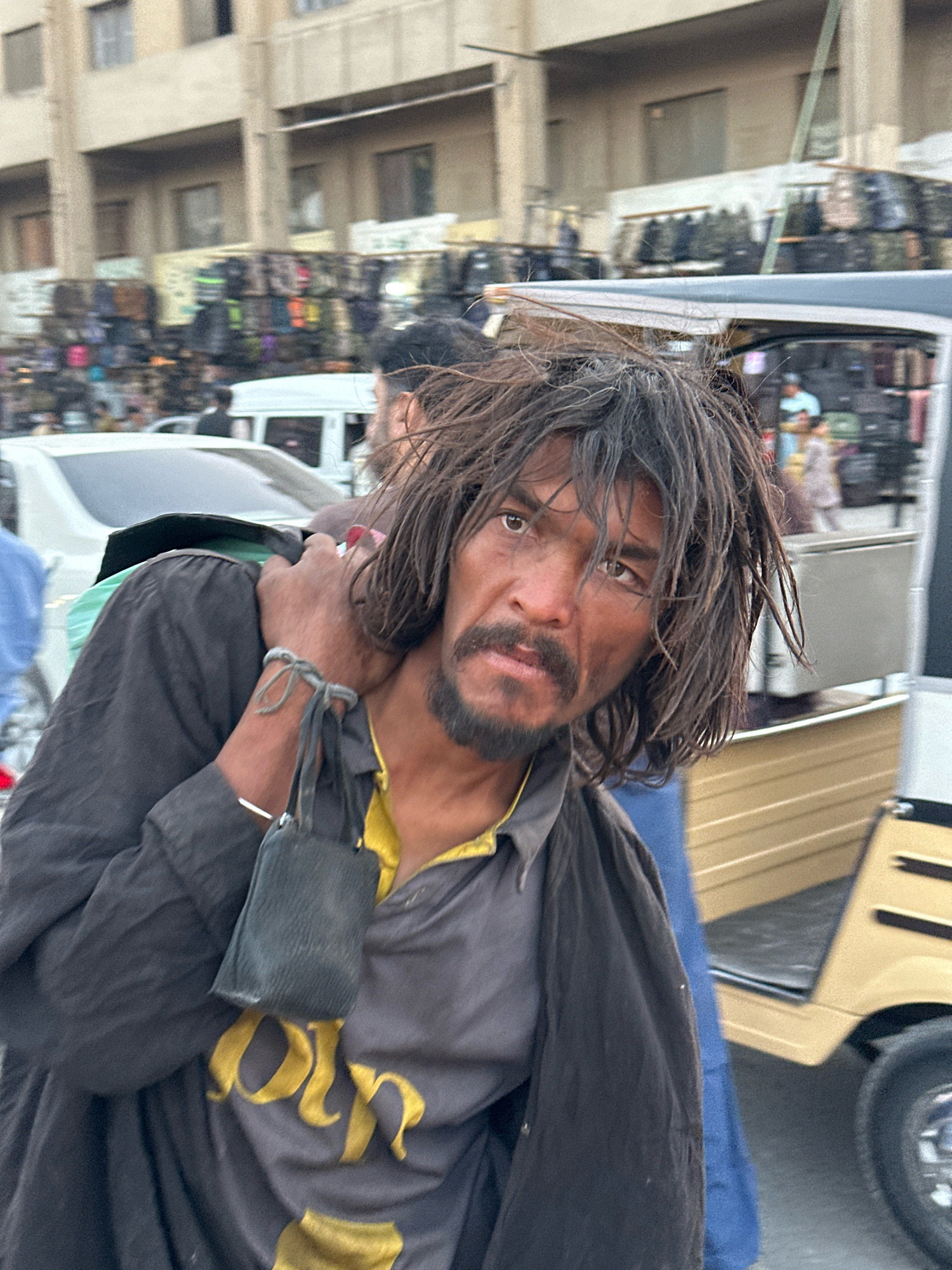 The image shows a close-up of a man with long, unkempt hair and a beard, wearing a dark jacket over a grey shirt with yellow lettering, and a yellow collar. He has a wristband with a small black pouch attached. The man is looking directly at the camera. In the background, there are buildings, market stalls displaying various items, vehicles including a white car and a yellow auto-rickshaw, and other people. The scene appears to be a busy urban environment. The image is likely taken in Quetta City Tehsil, Pakistan, given the architectural style of the buildings and the type of vehicles visible.