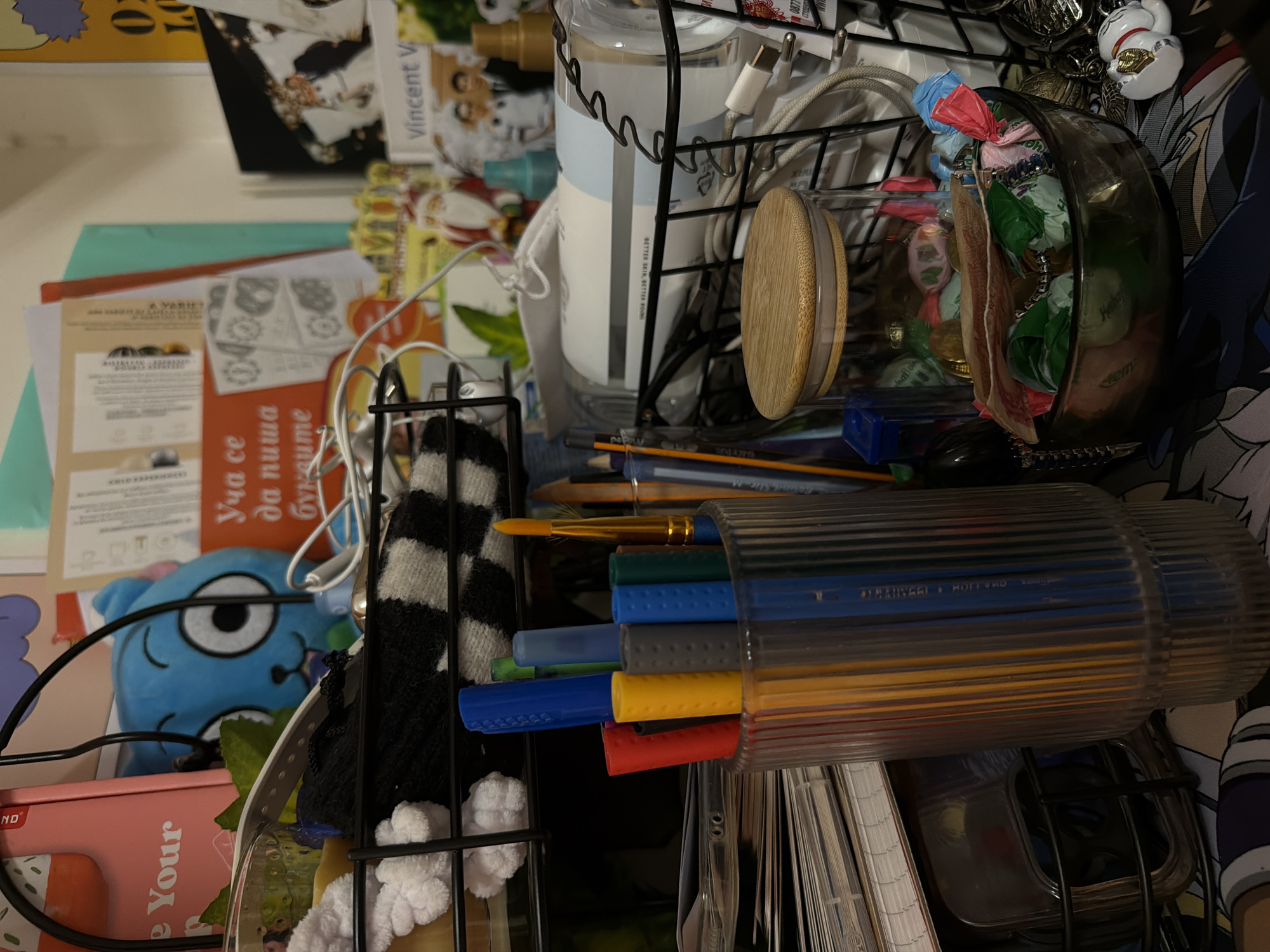 The image depicts a cluttered desk or shelf arrangement. In the foreground, a clear ribbed glass container holds an assortment of colorful markers, including red, yellow, gray, blue, and green. Behind this, a black wire basket contains various items, including a white object with a textured surface, a black and white striped item, and what appears to be a roll of paper or fabric. Further back, a glass jar with a wooden lid is filled with candies wrapped in colorful paper. A small dark bowl also contains candies and other small objects. Various papers, magazines, and other assorted items are stacked and arranged around the desk. A small blue plush toy with large eyes is visible in the background, partially obscured by other objects. The lighting suggests an indoor setting, possibly artificial. There is no discernible location information within the image that would indicate Sofia, Bulgaria.