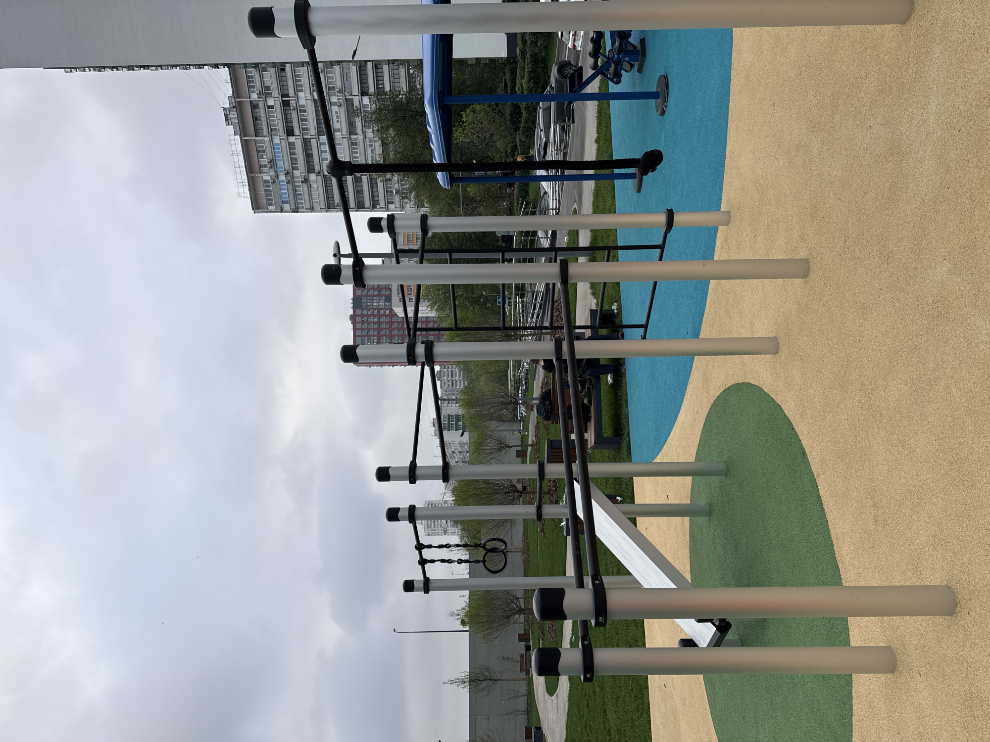 The image depicts an outdoor fitness area in Moscow, Russia. The area features parallel bars, gymnastic rings, and pull-up bars. The ground beneath the equipment is covered with colored rubber padding in shades of blue, green, and tan. In the background, apartment buildings and trees are visible. The sky is overcast. There are people in the background near the park benches.