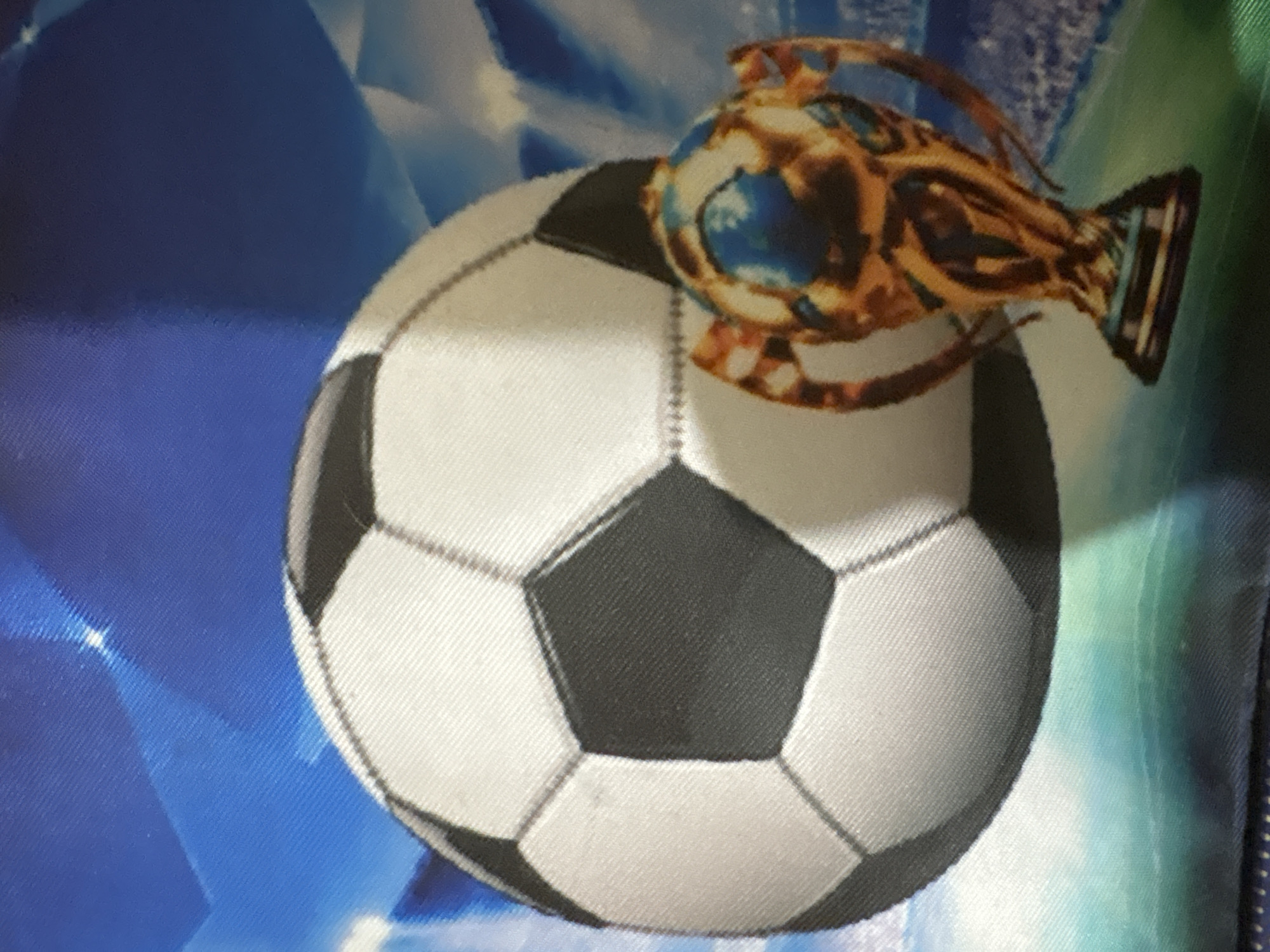 A close-up, slightly blurry, overhead shot features a soccer ball and a trophy. The soccer ball, mostly white with black hexagonal panels, is positioned on the left side of the frame, taking up a significant portion of the image. To its right and slightly in front, a golden trophy with a dark base is visible. The trophy has intricate detailing and appears to be a representation of a sports award. The background is a stylized depiction of a soccer stadium, with bright blue and green elements suggesting stadium lights and a field. The image appears to be printed on fabric, given the visible texture. The location of this image in Ashvem, India, cannot be determined from the visual information provided.