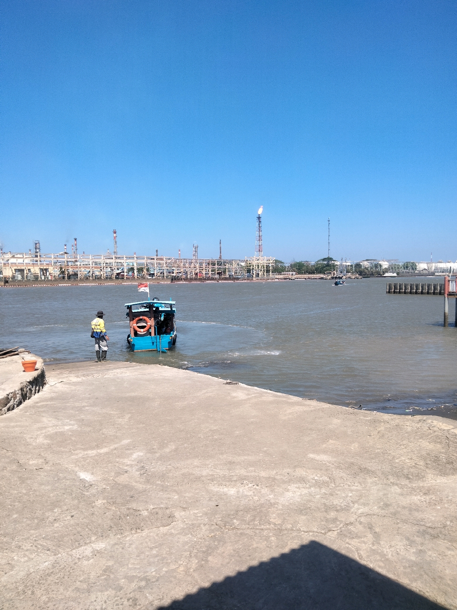 This is a vertical, eye-level, wide shot of a coastal industrial scene in Cilacap, Indonesia, under a clear blue sky. In the foreground, a concrete ramp slopes down to the water. A person in a yellow vest, gray pants, and a black hat stands on the ramp, facing a small blue boat. The boat has a life preserver hanging off its side and an Indonesian flag on its stern. The water is murky and appears calm. In the background, a large industrial complex with numerous pipes, towers, and buildings stretches across the horizon, with a flare stack emitting a flame. A few other boats are visible in the distance on the water. The overall atmosphere is one of industrial activity and a working port.