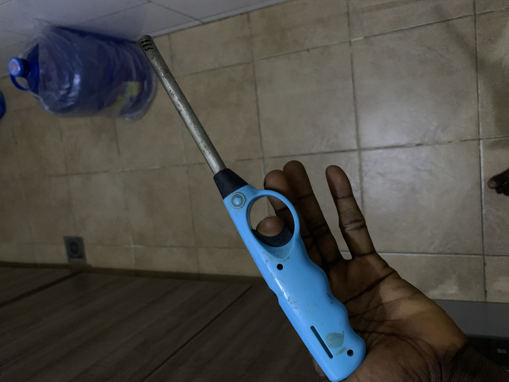 A dark-skinned hand holds a light blue, long-stemmed utility lighter in the foreground. The lighter features a metallic stem, a blue plastic handle with a circular finger loop, and a small, light-colored button near the top of the handle. The plastic handle shows signs of wear and scratches.
In the background, two large, translucent blue plastic water bottles are visible. They are partially encased in clear plastic bags, and one bottle has a blue cap. The upper portion of the background consists of a light brown or beige tiled floor with visible grout lines. Below this, the floor transitions to a darker, horizontal wooden or laminate surface. The scene is moderately lit, suggesting an indoor setting, likely a kitchen or utility area. No discernible text is visible on any objects. The overall impression is a domestic environment in Gwarinpa, Nigeria.