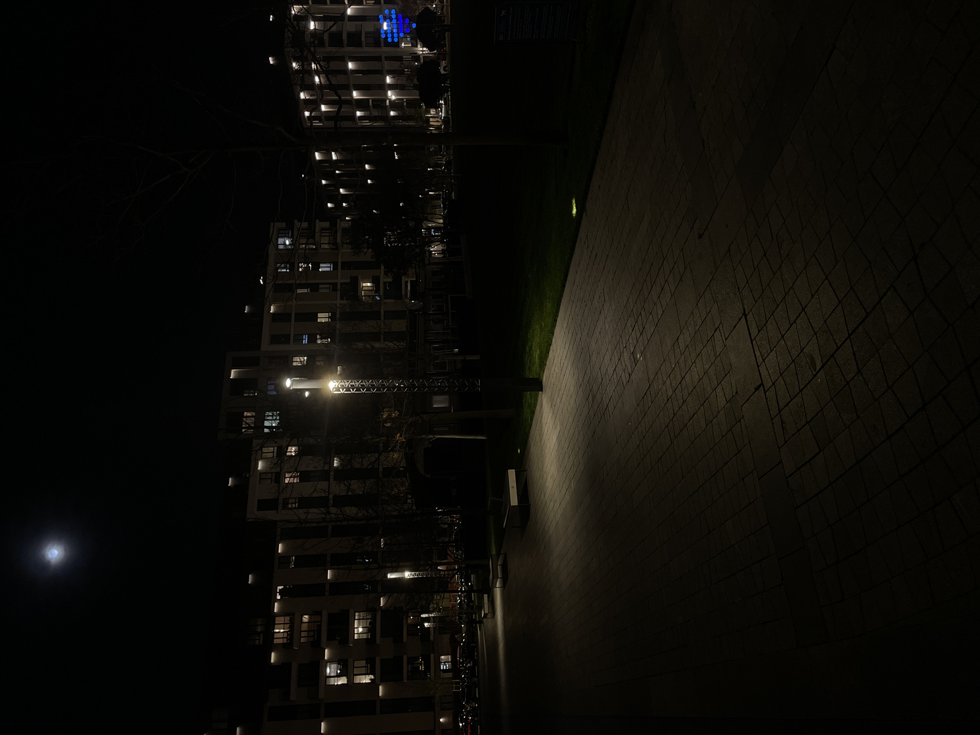 The image shows a nighttime scene in Belgrade, Serbia, with a view of modern apartment buildings illuminated by interior and streetlights, contrasting against the dark sky. The moon is visible to the left of the frame. A pedestrian walkway occupies the right side of the frame, leading away into darkness. The scene appears to be in a newly developed residential area.