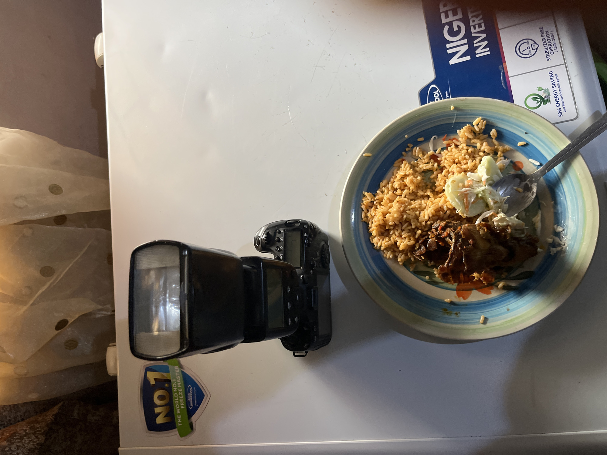 A digital camera with a flash attached is positioned on a white surface, possibly a refrigerator, which has a "NO.1 FREEZE MASTER" sticker and a "NIGER INVERTER" label visible. In the foreground, a plate of food, appearing to be rice with meat and some salad, is placed on the same surface. The lighting suggests the image was taken indoors with artificial light. The overall scene conveys a domestic setting, though the specific location in Bukuru, Nigeria cannot be determined from the visual information alone.
